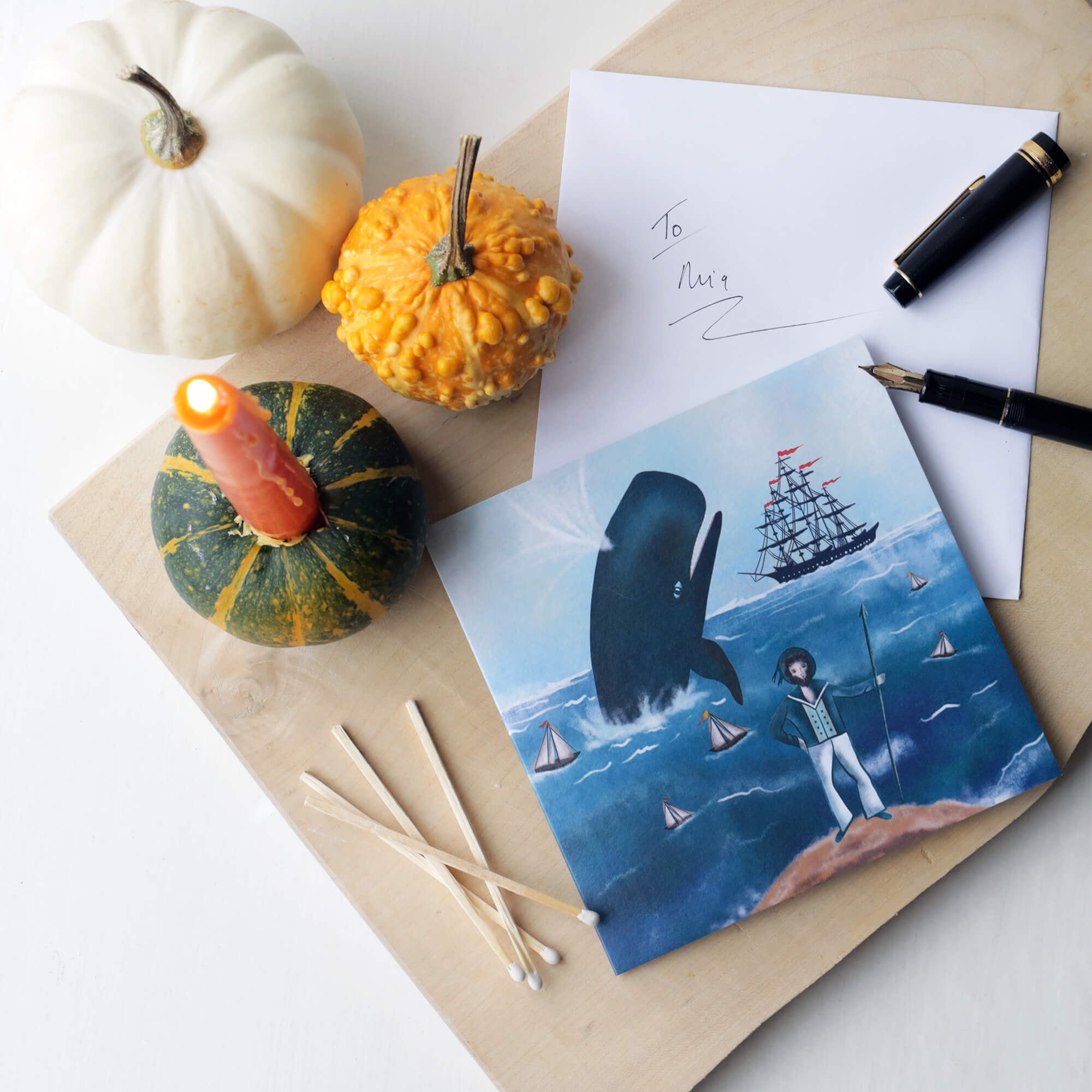 A flat lay display of a nautical-themed greeting card featuring a sailor, a breaching whale, and a tall ship. The card is placed on a wooden surface with autumnal decorations, including a white pumpkin, a yellow gourd, and a lit candle inserted into a small green pumpkin. Nearby are matches, an envelope addressed 'To Mia,' and a fountain pen.