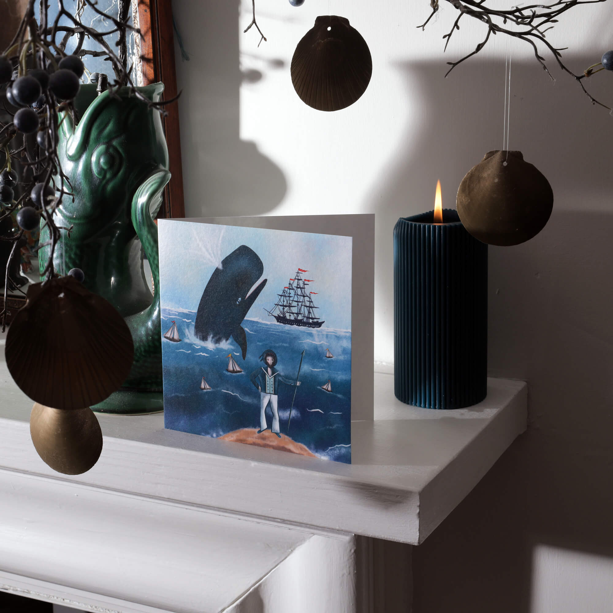 A nautical-themed greeting card featuring a sailor, a breaching whale, and a tall ship displayed on a mantelpiece. The setup includes coastal decor, such as hanging seashell ornaments, a dark green ceramic vase, and a lit blue candle. Soft natural light enhances the maritime atmosphere, casting gentle shadows across the scene.