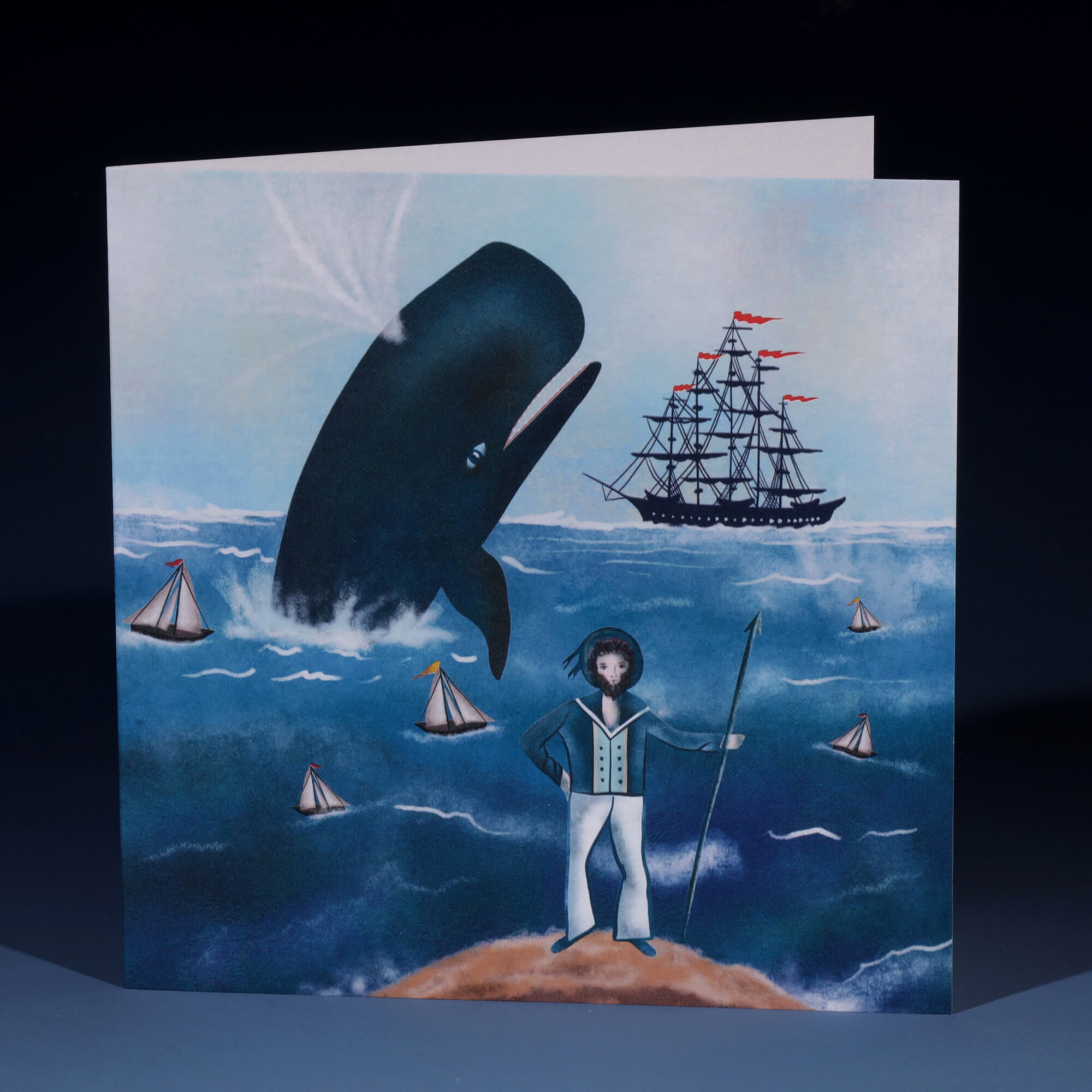 A nautical-themed greeting card featuring an illustration of a large whale breaching the ocean, a sailor standing on a rock holding a spear, and a tall ship with red flags in the background. Several small sailboats are scattered across the water, creating a whimsical, adventurous scene in soft blue tones.