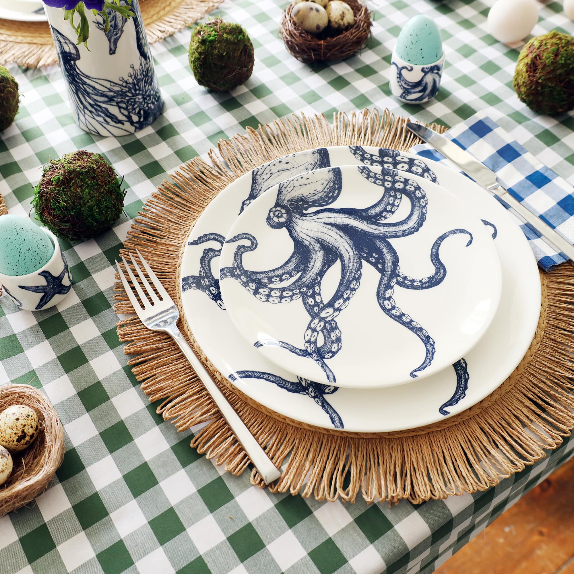 2 white plates on top of each other each with a navy octopus on them. They are on top of a raffia placemat on a green checked tablecloth with balls of moss and eggs for easter on the table.