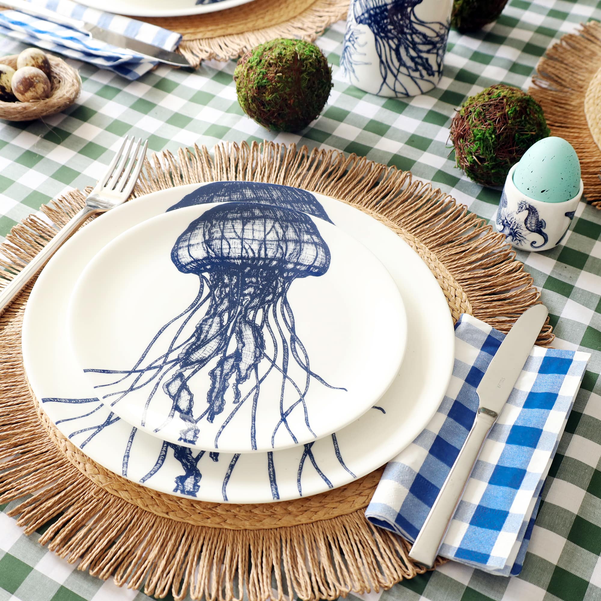 White bone china dinner plate with side plate sitting on top. Both plates have a navy blue illustration of a jellyfish on. They are set as a place setting on a raffia placemat with blue gingham napkin and green gingham tablecloth. the table is set for Easter with eggs in egg cups and moss balls.