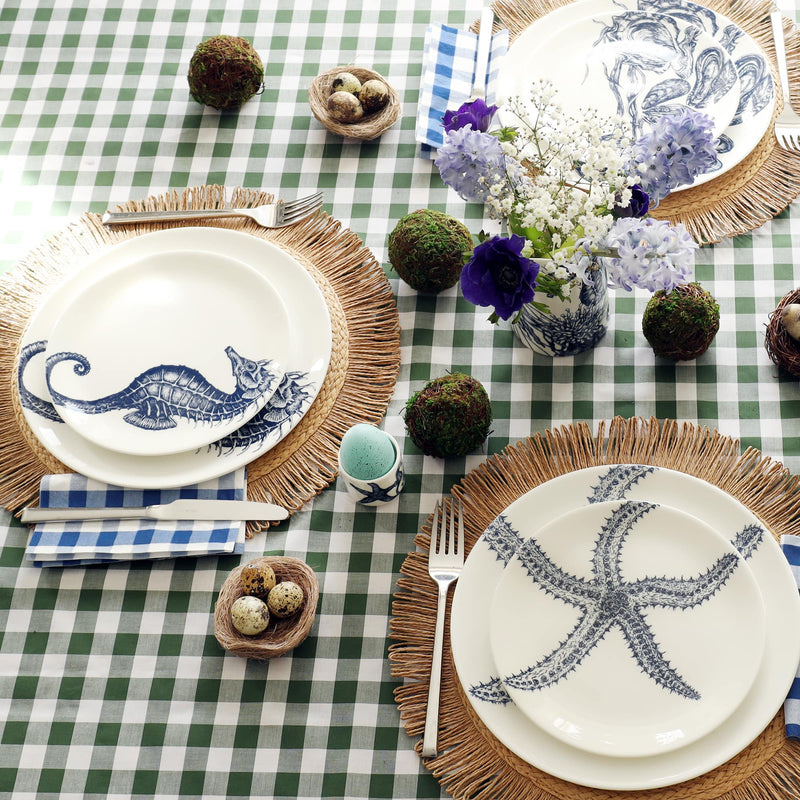 Bone China White dinner plate with side plate in top with hand drawn illustrations a starfish, seahorse and mussels and oysters in navy. These three place settings are sitting on a raffia placemat and set with a knife and fork each side. The table is laid with a green gingham tablecloth and there are eggs and moss balls decorating the table for Easter.