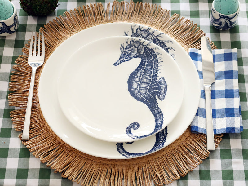 Table setting with 2 white bone china plates, one dinner one side plate sitting on top each with a beautifully illustrated seahorse in navy blue. They are on top of a raffia placemat and green gingham tablecloth. 