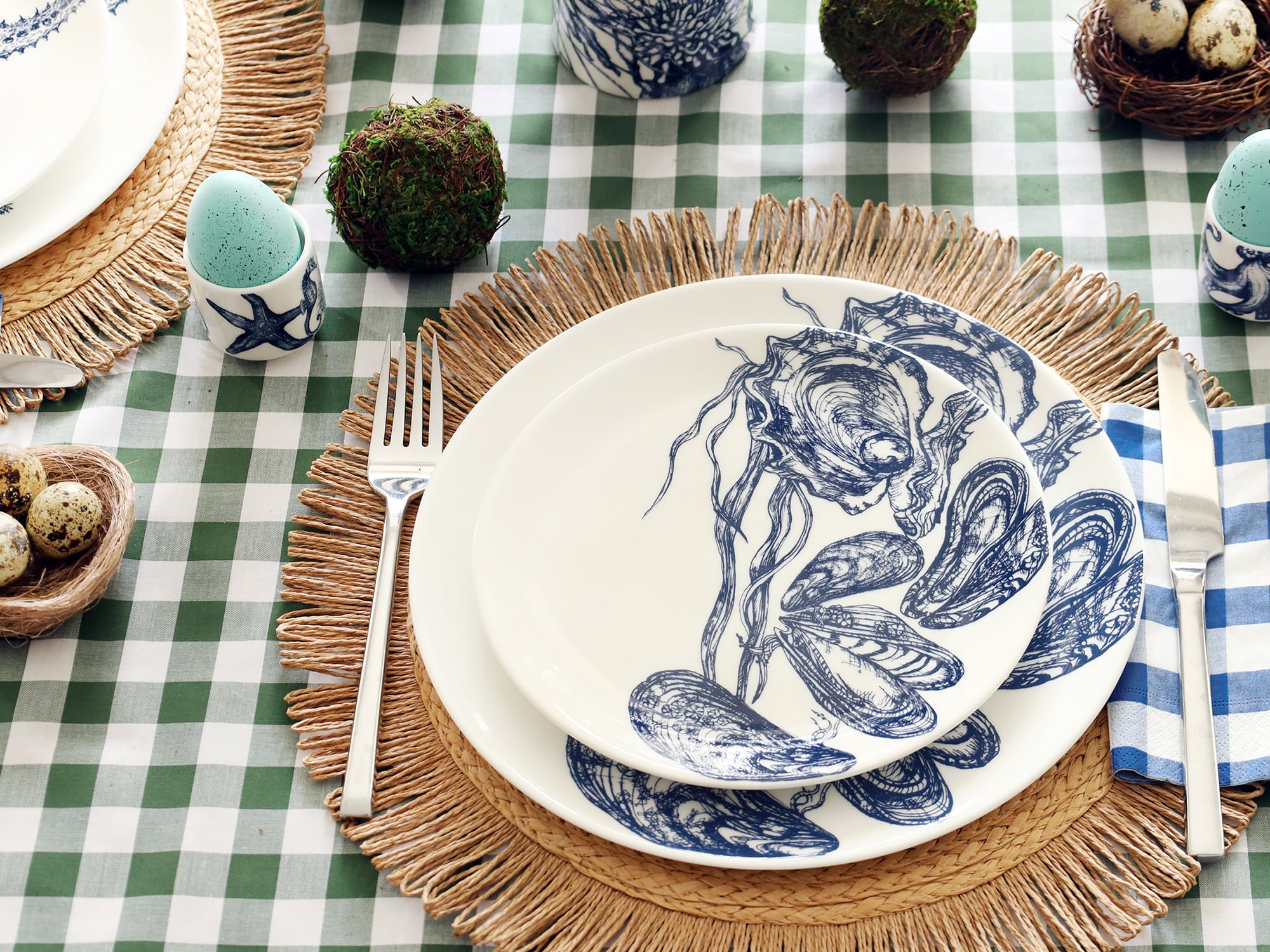 White bone china dinner plate with side plate sitting on top. Both plates have a navy blue illustration of mussels & oysters on. They are set as a place setting on a raffia placemat with blue gingham napkin and green gingham tablecloth. the table is set for Easter with eggs in egg cups and moss balls.