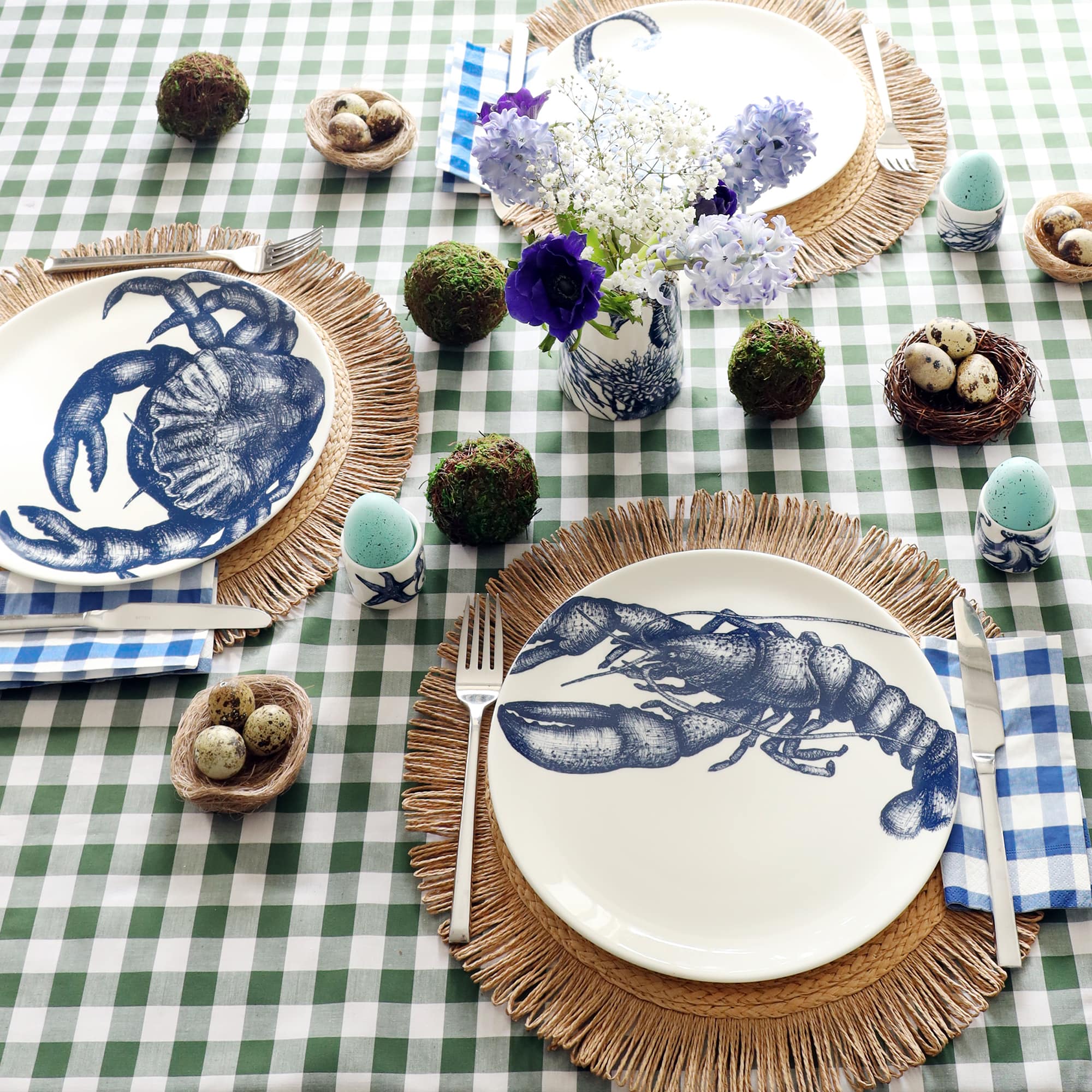 Easter table setting with blue and white china with sea creatures on. In the foreground is a plate with a navy lobster illustration, sitting on a raffia placemat and green gingham tablecloth. There are moss balls, light blue eggs and small nests with quails eggs in decorating the table.