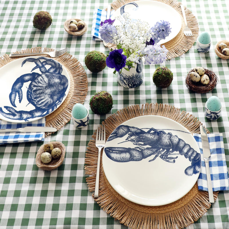 Easter table setting with blue and white china with sea creatures on. In the foreground is a plate with a navy lobster illustration, sitting on a raffia placemat and green gingham tablecloth. There are moss balls, light blue eggs and small nests with quails eggs in decorating the table.