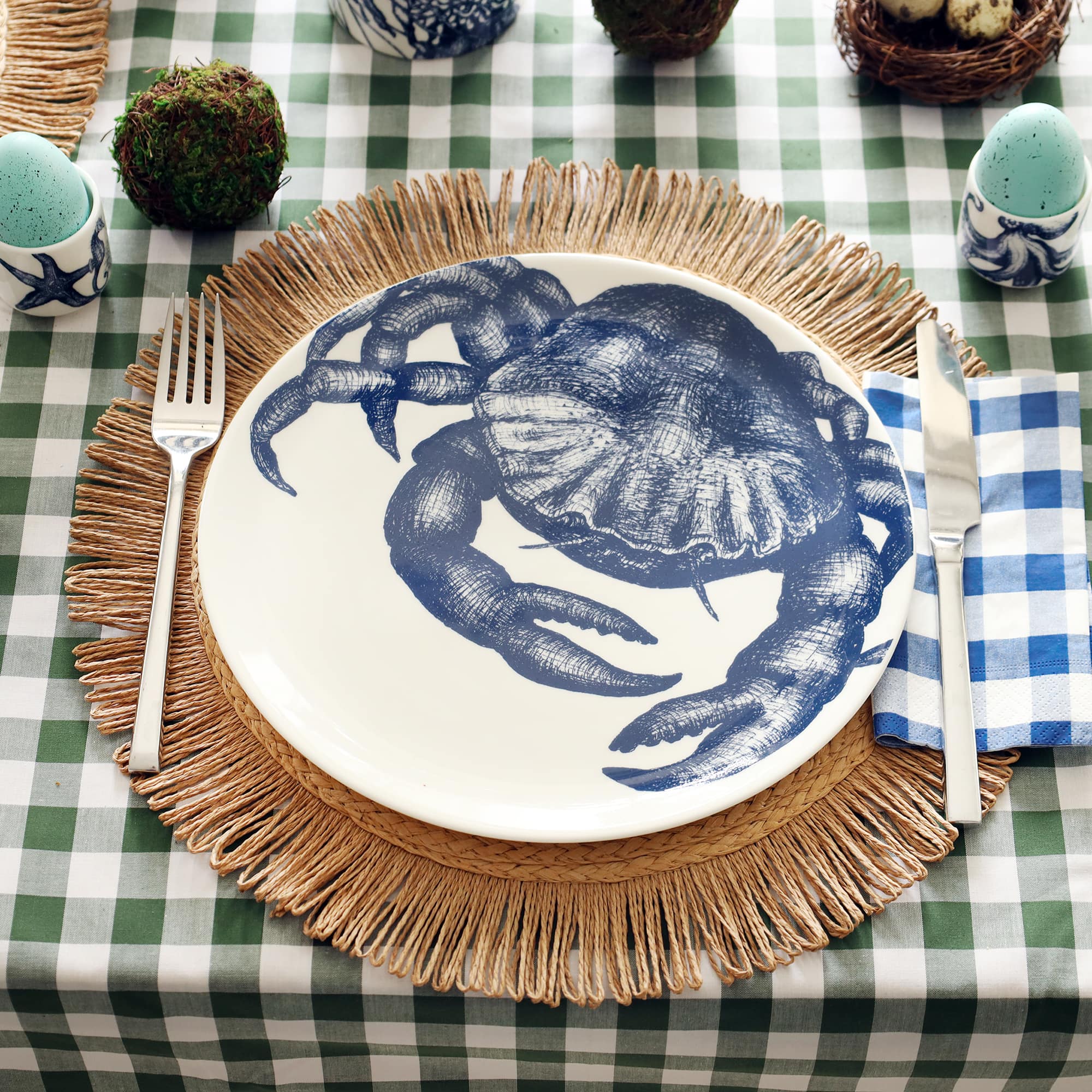 White bone china plate with navy crab illustration sitting on a raffia placemat and set for dinner with knife and fork either side. The table is laid for Easter with blue eggs in egg cups and moss ball to decoarate.