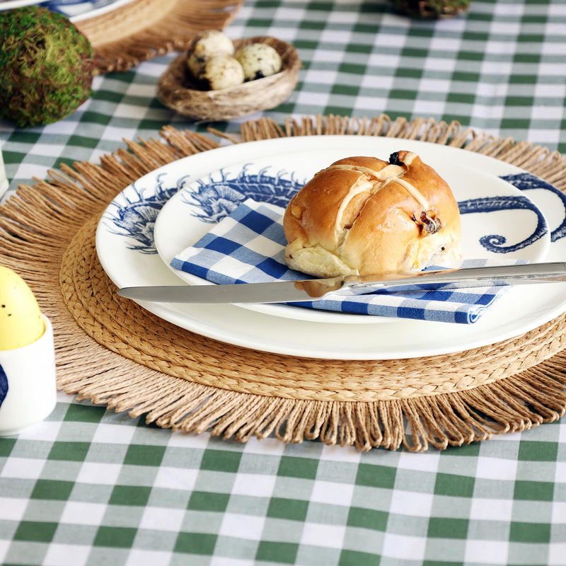 2 white plates on top of each other, each with a navy seahorse illustration on them. There is a  blue gingham napkin folded on the plate a hot cross bun and knife. The plate is on a raffia placemate and this is on a green gingham tablecloth.  