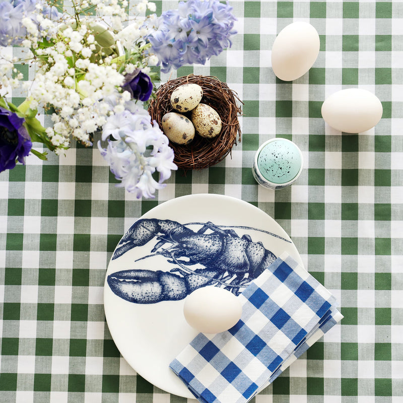 White plate with navy blue lobster decoration with folded blue gingham napkin and duck egg on it. This is on a green gingham tablecloth with a small nest and quails eggs, hyacinths and anemones and more eggs.