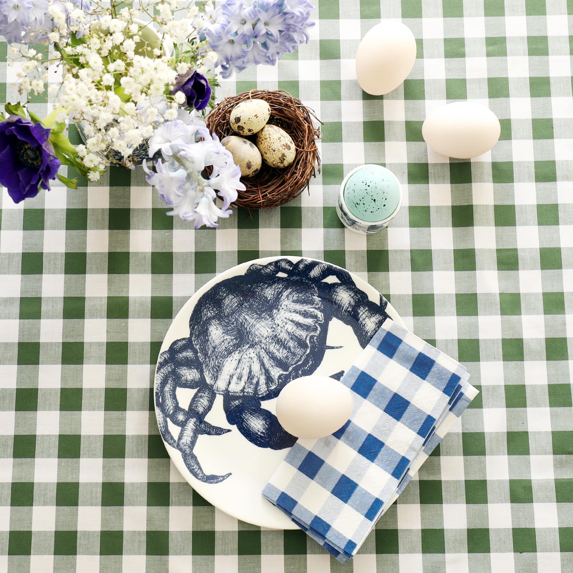 White plate with navy blue crab decoration with folded blue gingham napkin and duck egg on it. This is on a green gingham tablecloth with a small nest and quails eggs, hyacinths and anemones and more eggs.