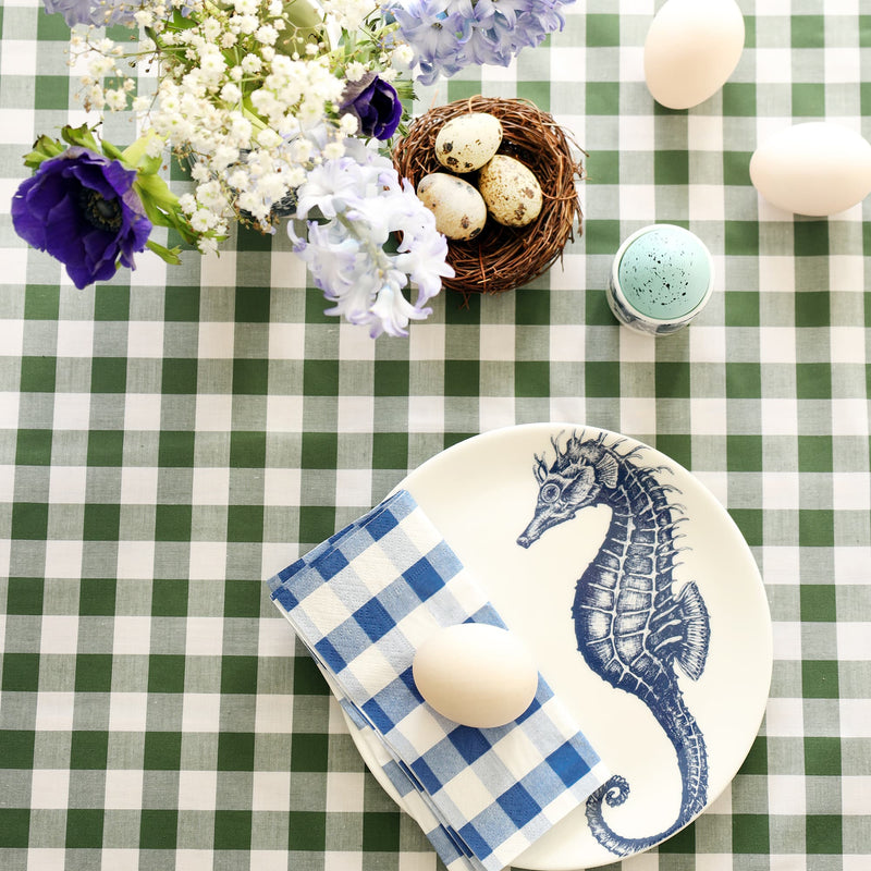 White plate with navy blue seahorse decoration with folded blue gingham napkin and duck egg on it. This is on a green gingham tablecloth with a small nest and quails eggs, hyacinths and anemones and more eggs.