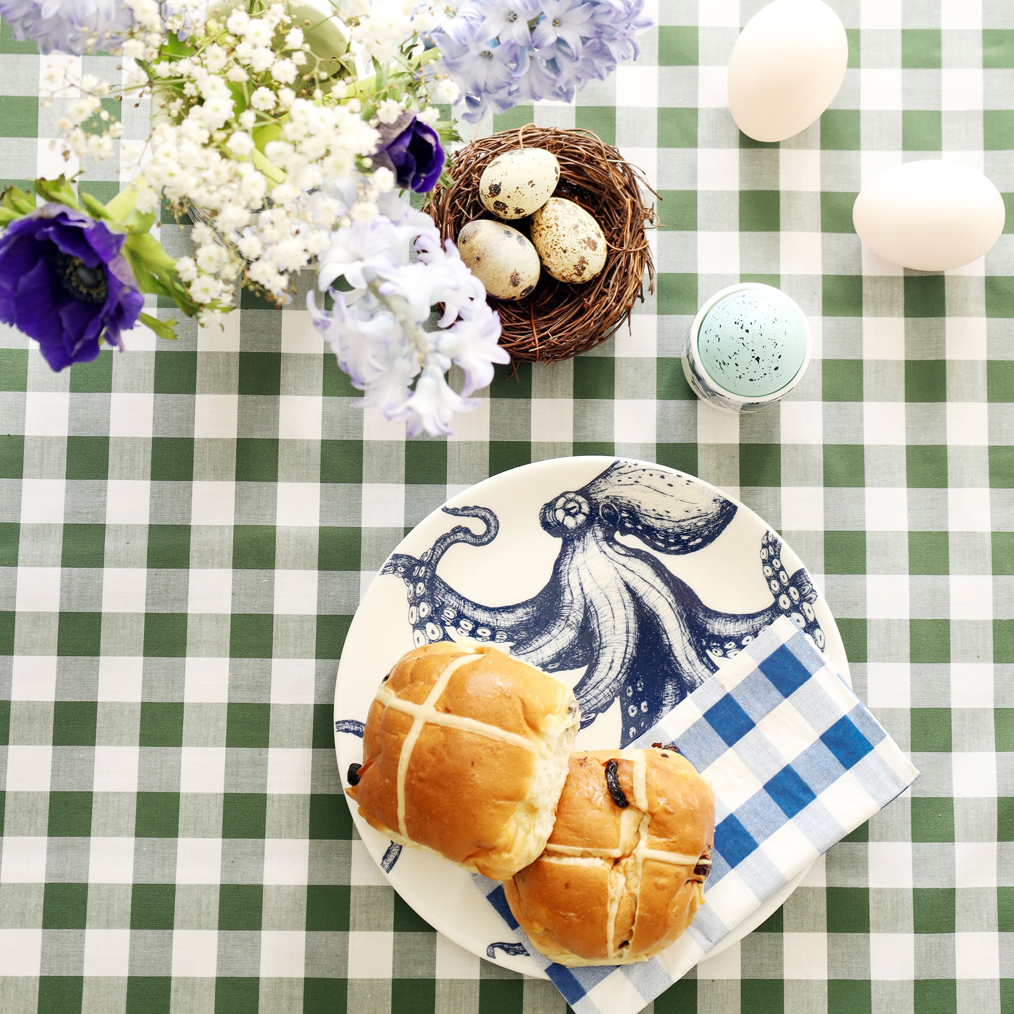 White plate with navy octopus illustration it with 2 hot cross buns and a blue checked napkin. There are flowers and eggs decorating the green checked tablecloth.