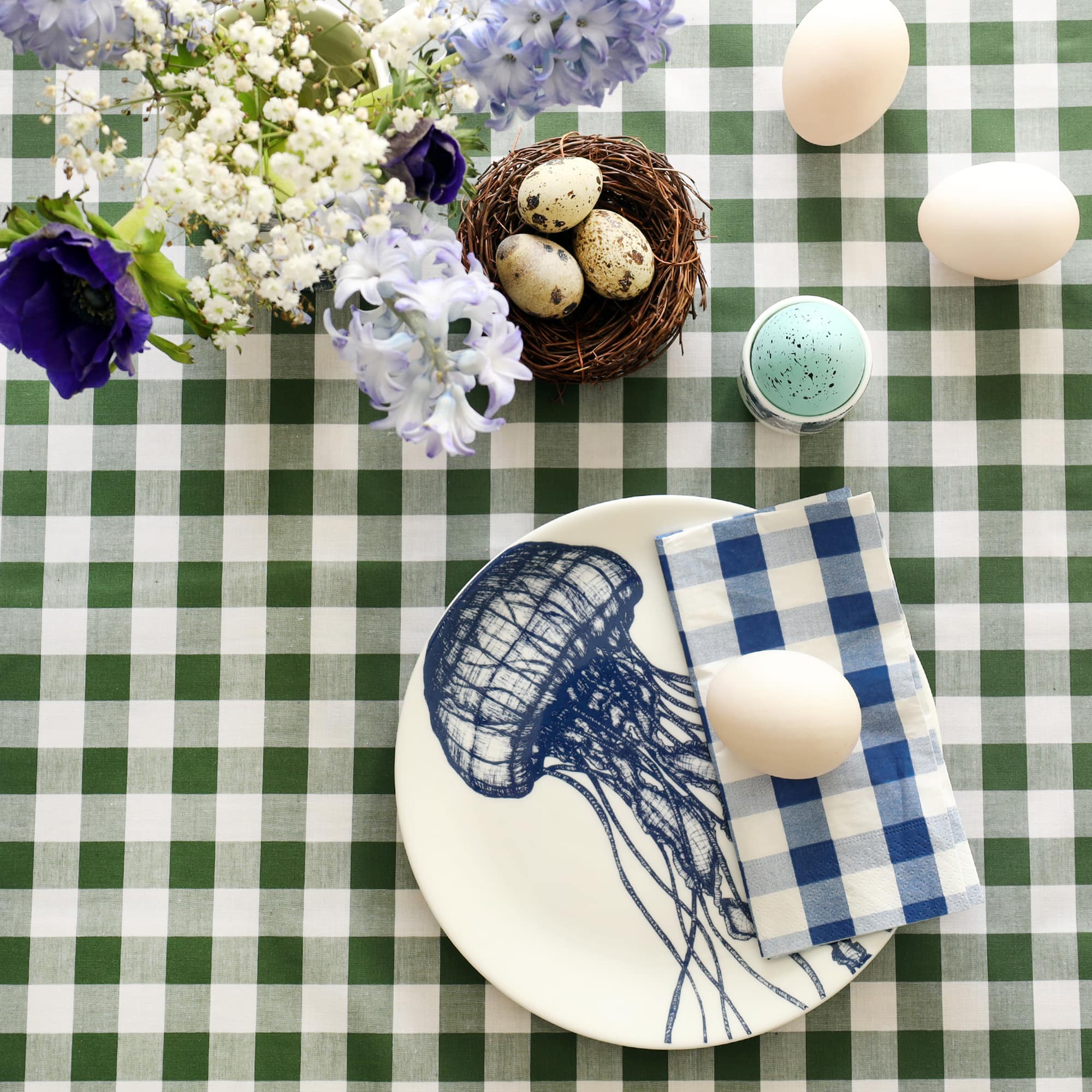 Bone china plate with illustration of jellyfish in navy. There is a fold blue gingham napkin and duck egg on the plate. The plate is on a green gingham tablecloth with a small nest and quails eggs and there are hyacinths and anemones in a jug.