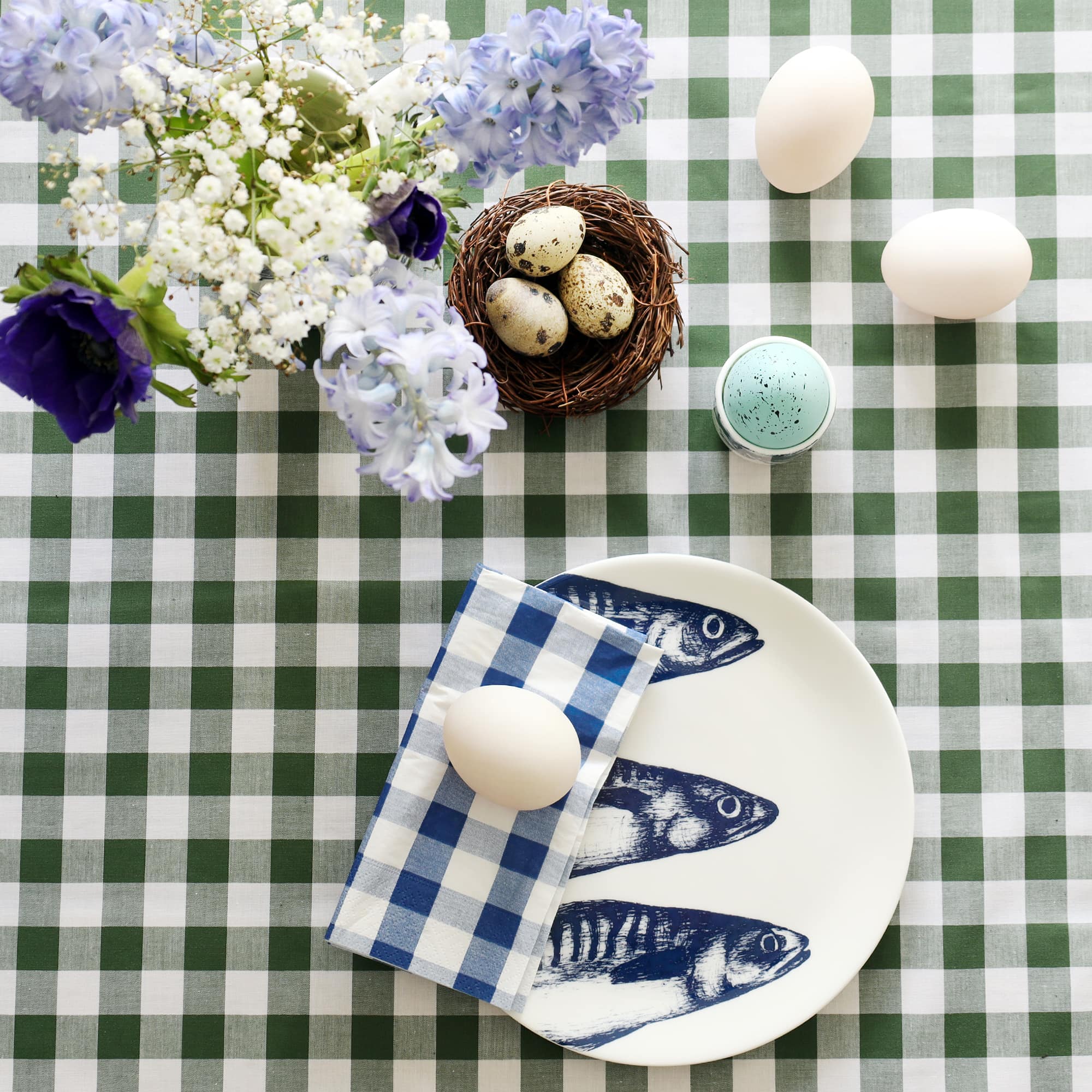White bone china plate with 3 mackerel heads illustration in navy on it. There is a folded blue gingham napkin on the plate with a duck egg. The table is laid in a green gingham tablecloth and there are more eggs, a small nest with quails eggs and a jug with hyacinths and anemones in.