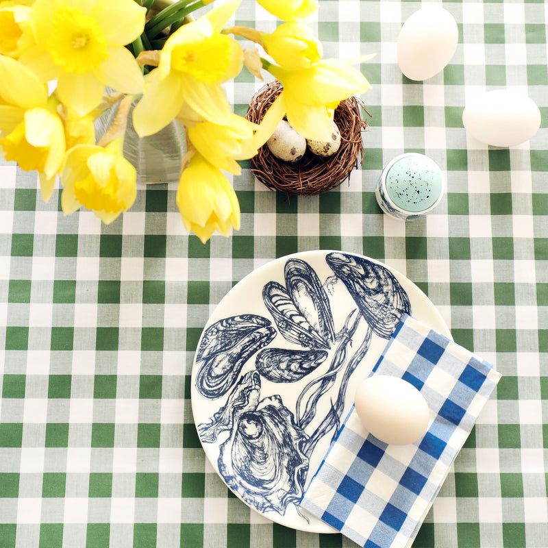 Bone China White dinner  plate with hand drawn illustrations of mussels and oysters in navy. There is a folded blue gingham napkin with a duck egg on the plate. The table is laid for Easter with a green gingham tablecloth, eggs and daffodils.