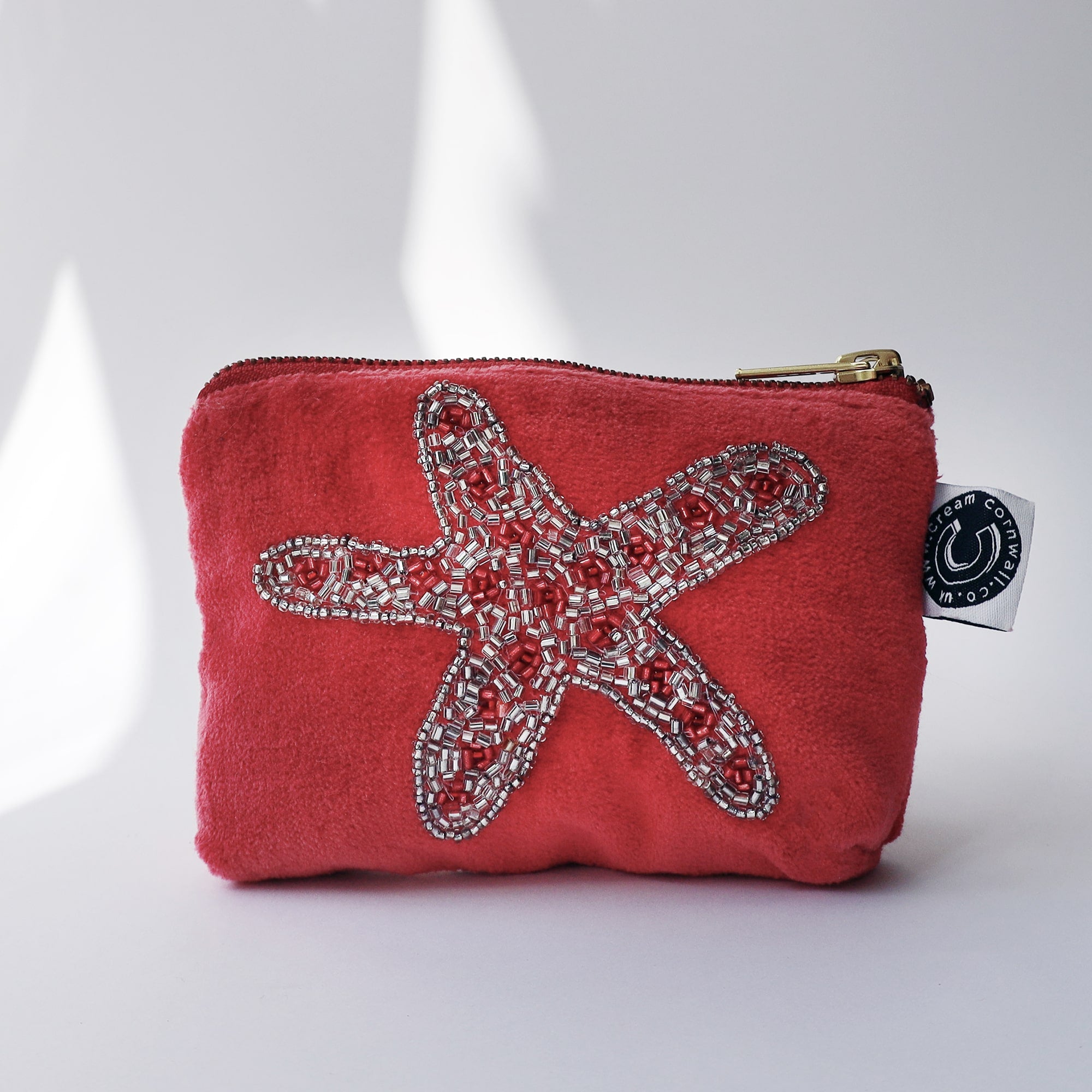 Pink velvet coin purse with a beaded starfish on the front sitting on white background with the sun casting shadows across it.