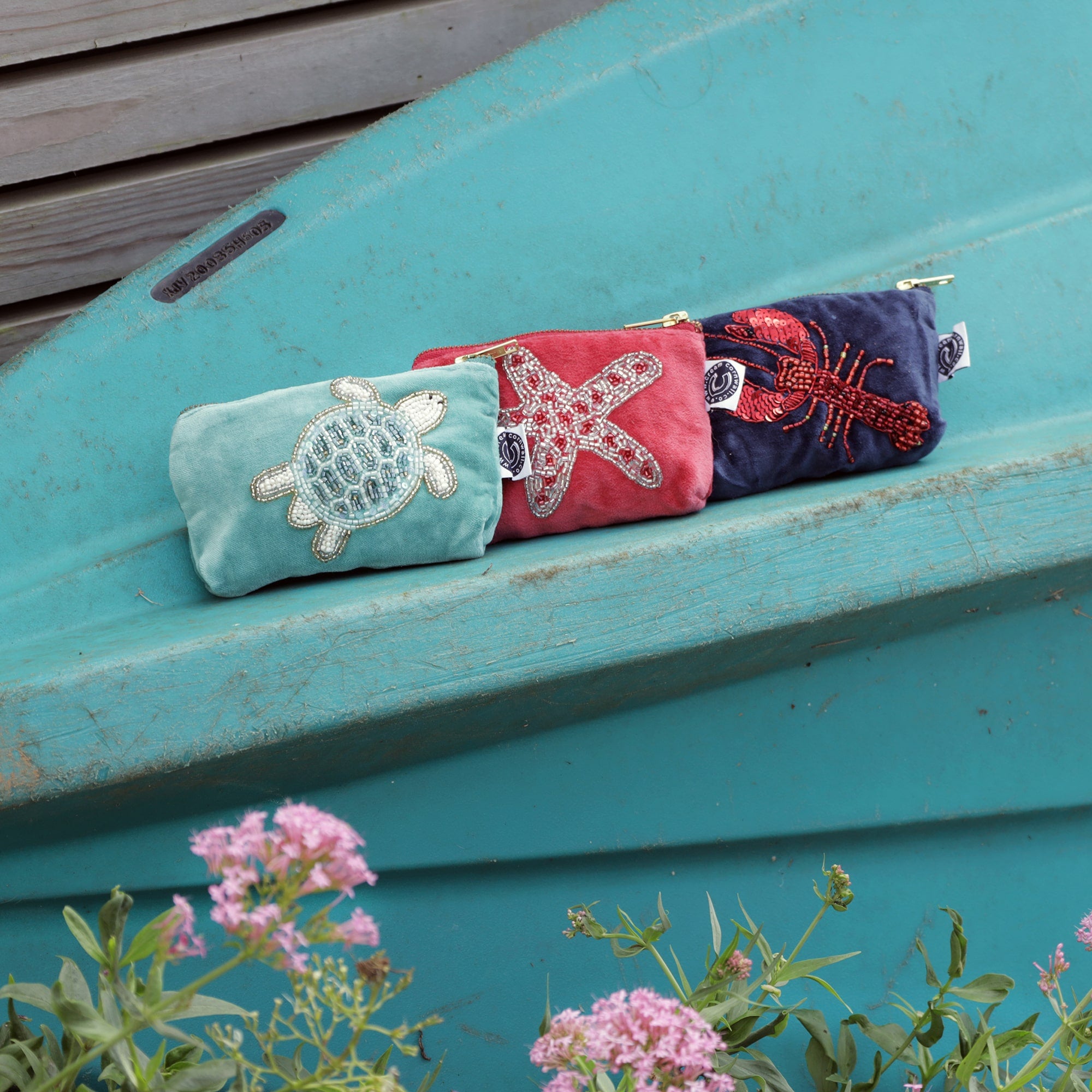 Three colorful coin purses are lined up side by side on a weathered turquoise surface, likely a boat or outdoor bench. The left pouch is light green with a beaded sea turtle design. The middle pouch is coral pink with a beaded starfish. The right pouch is navy blue with a red sequined lobster. In the foreground, pink flowers slightly blur, adding a touch of nature to the coastal-themed scene.