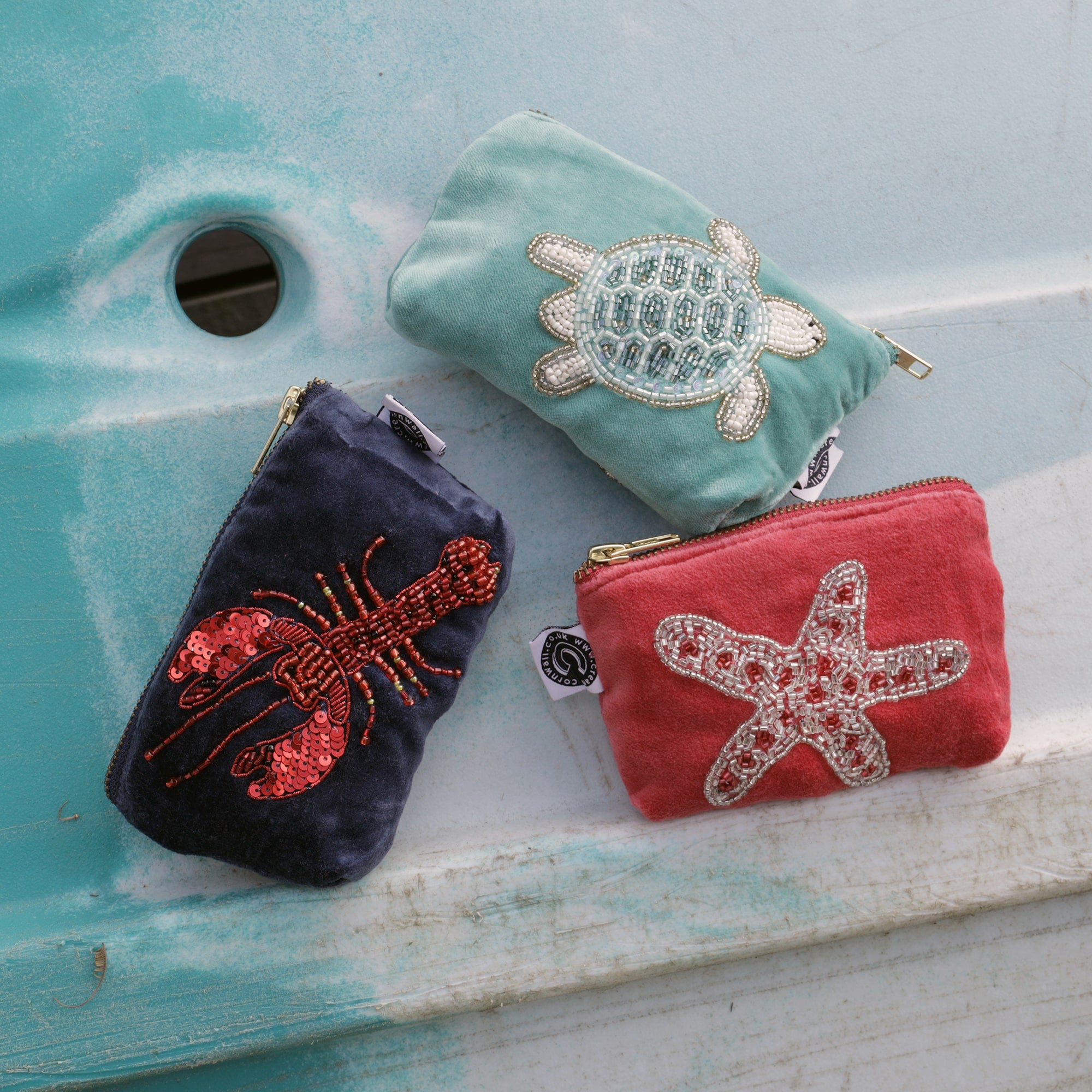 Three small fabric coin purses are displayed against a light blue background. Each pouch is uniquely designed with sea-themed embroidery and sequins. The top pouch is light green with a beaded sea turtle. The middle pouch is navy blue with a red sequined lobster. The bottom pouch is coral pink with a beaded starfish. All pouches have gold zippers and small white tags attached.