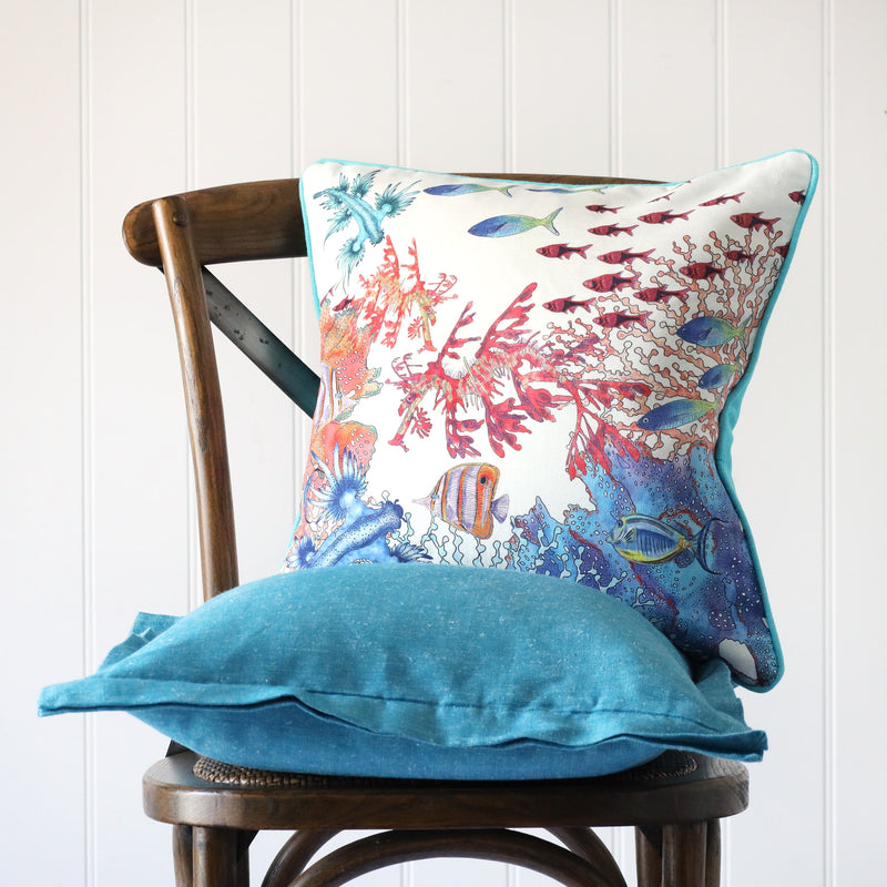 white cushion with brightly coloured illustration of a tropical underwater scene on the front, placed on a turquoise linen cushion on a wooden chair