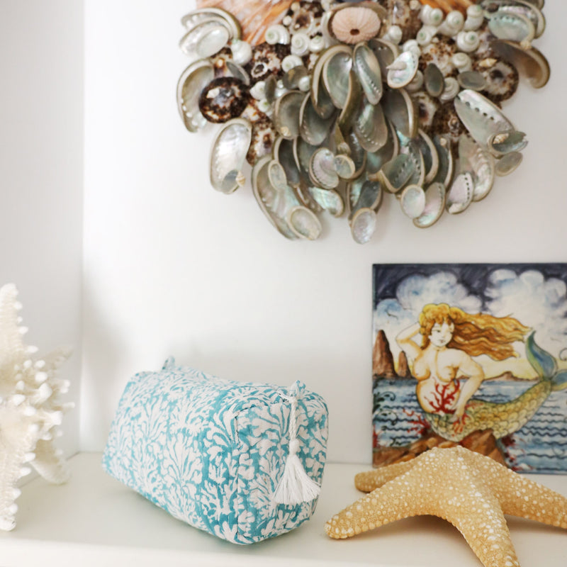 Tubular shaped cosmetic bag with Summer Skies Coraline design placed on a shelf with a starfish and a mermaid painting.On the wall in the background is a shell wall decoration