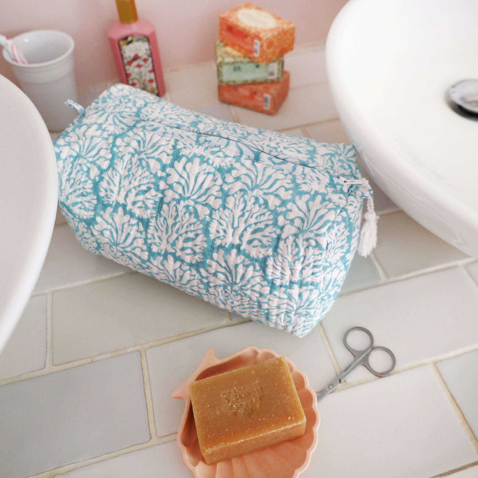 Tubular shaped cosmetic bag with Summer Skies Coraline  design on a tiled surface in between two sinks.In front is a shell with a handmade soap in, behind are wrapped soaps,toothbrush holder and Handcream