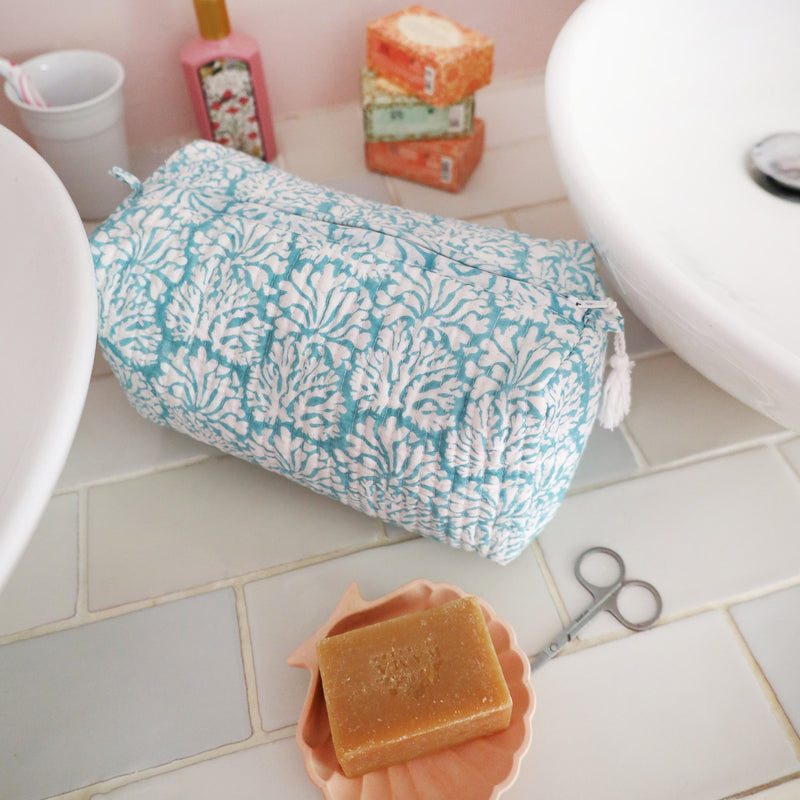Tubular shaped cosmetic bag with Summer Skies Coraline  design on a tiled surface in between two sinks.In front is a shell with a handmade soap in, behind are wrapped soaps,toothbrush holder and Handcream