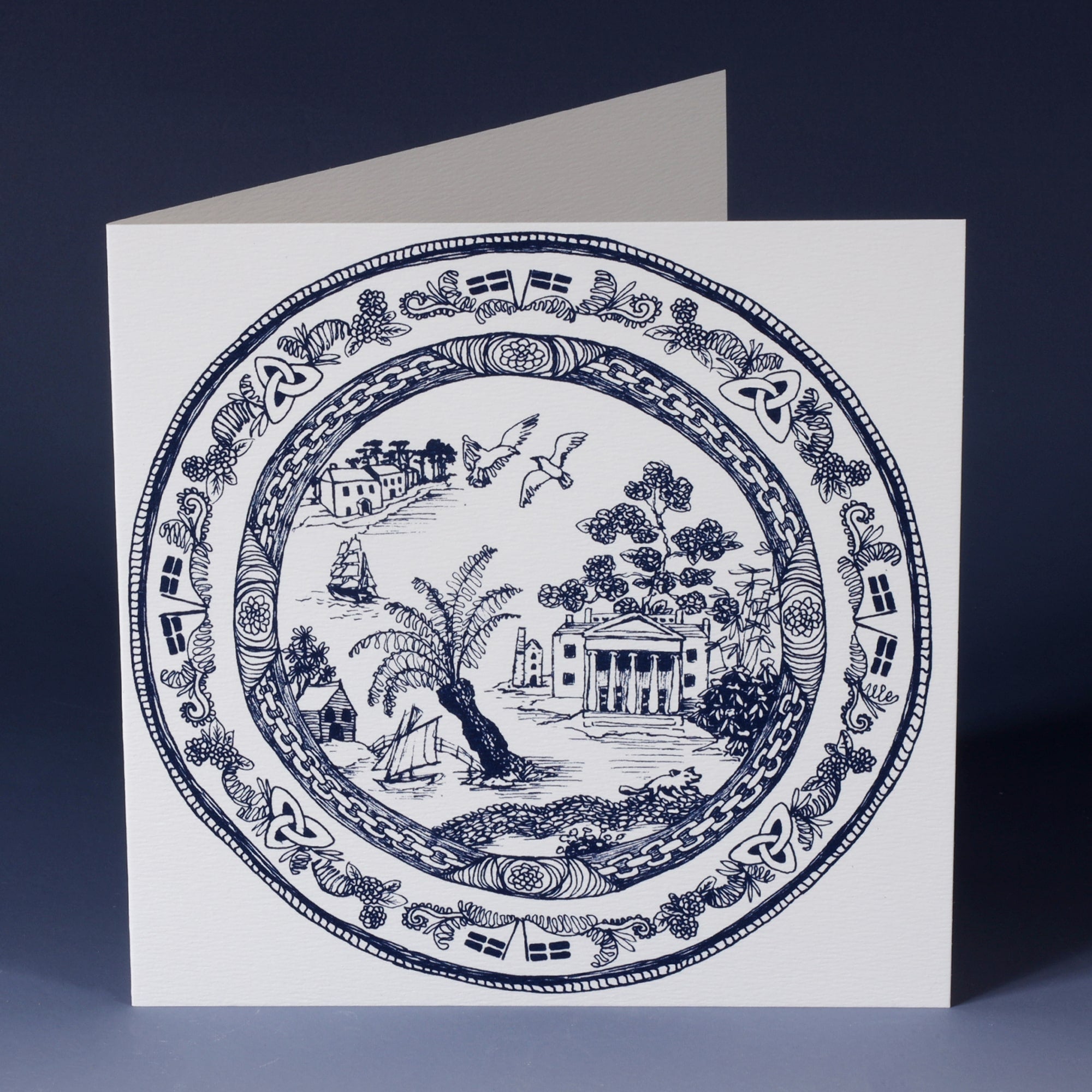 white greeting card with navy illustration of a cornish inspired willow pattern plate, with trelissick gardens, seagulls, tree ferns fishermens cottages and cornish flags 