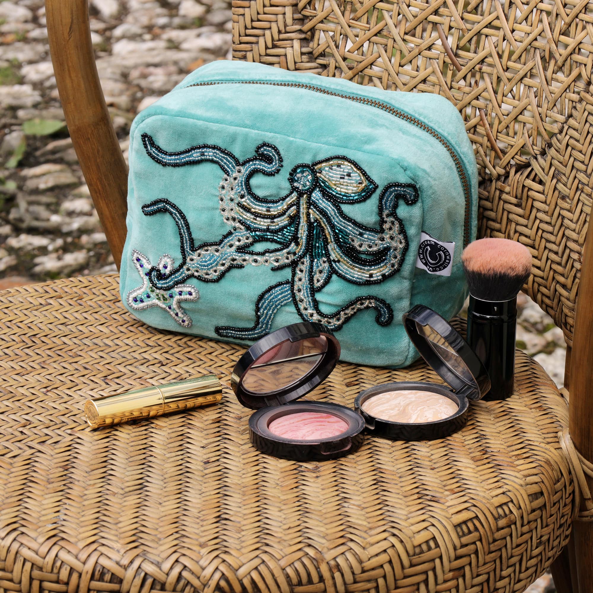 Beaded aqua velvet cosmetic bag with octopus and starfish decoration sitting on a wicker chair with some cosmetics on the foreground.