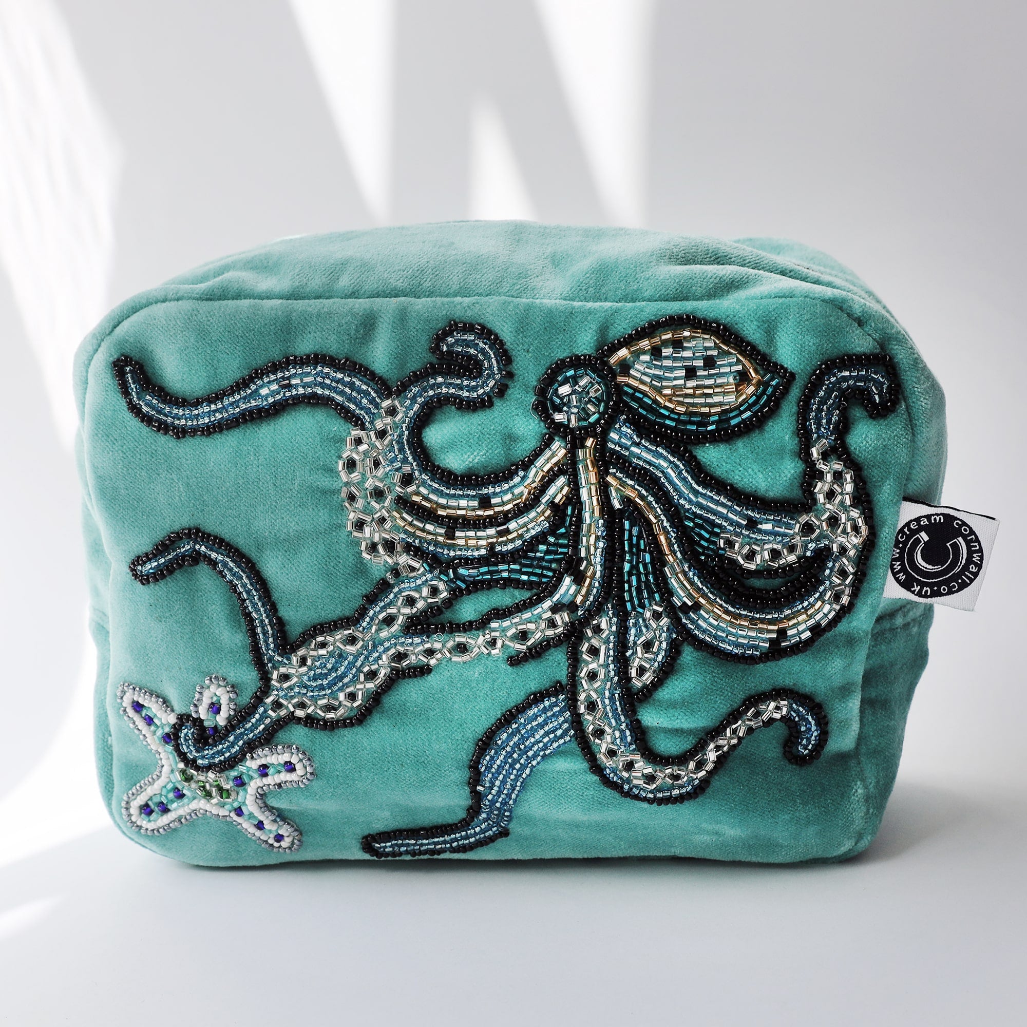 Beaded aqua velvet cosmetic bag with octopus and starfish decoration on the front on a white background with the sun casting shadows across it.