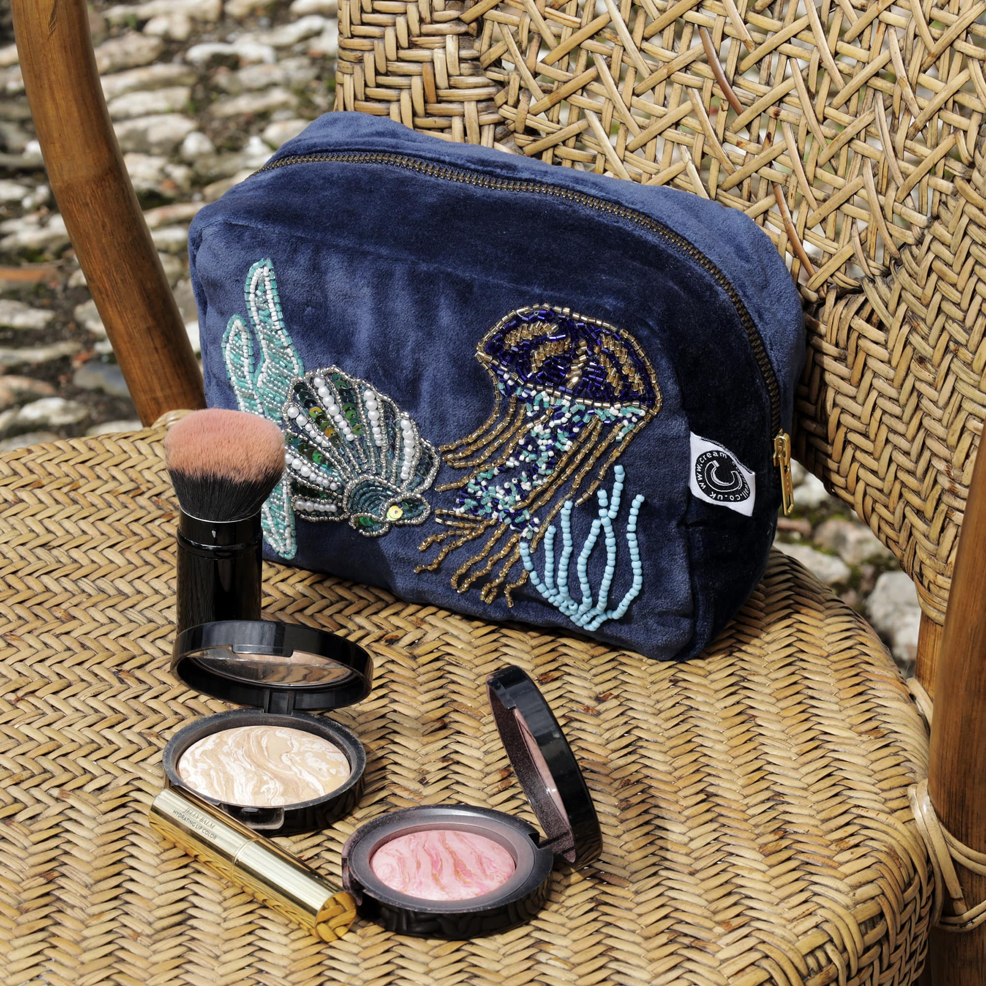 Beaded cosmetic bag in navy velvet with underwater scene of jellyfish, scallop shell and seaweed sitting on a wicker chair outside with cosmetics and brush in front of it.