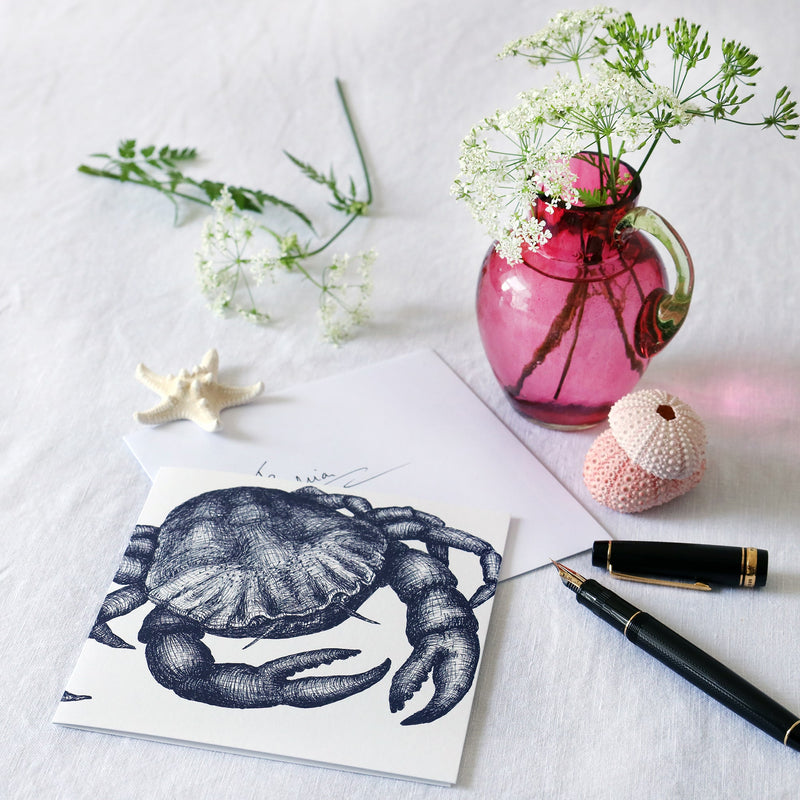 greeting card with navy illustrated crab on a white background lying on a white table cloth with a fountain pen, hand written envelope shells and a small cranberry glass jug with wild flowers in 