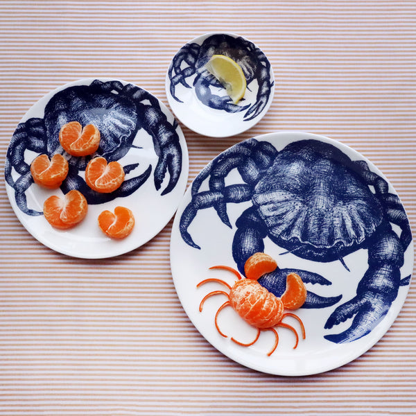 Three different sized white bone china plates with an illustrated navy blue crab on them. The largest plate has a crab made from a satsuma on it and the other 2 have a crab claws made from a satsuma and a slice of lemon on it. Both plates are shot from above on a striped yellow gold cloth.