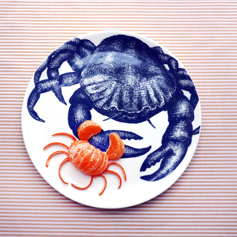 A white bone china plate with a navy illustrated crab on it. There is a smaller crab made from a satsuma on it and it is sitting on a golden yellow striped cloth.