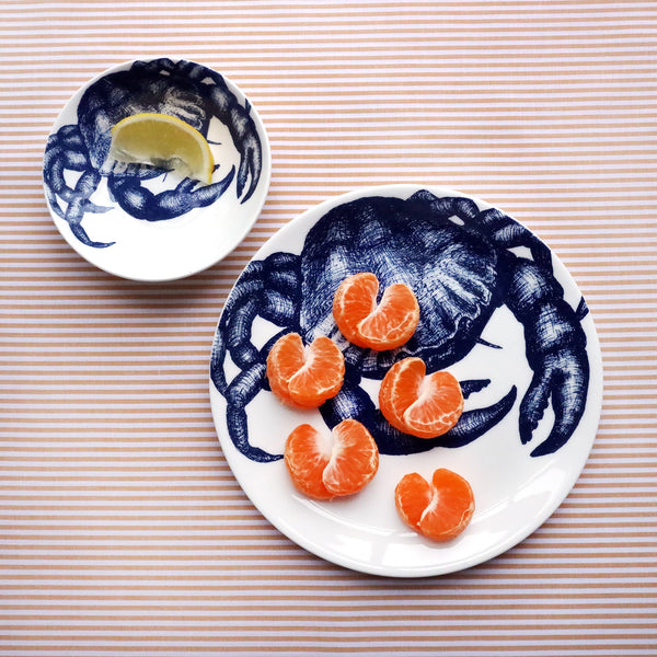 Two different sized white bone china plates with an illustrated navy blue crab on them. The largest plate has a crab made from a satsuma on it and the other one has a slice of lemon and a slice of lemon on it. Both plates are shot from above on a striped yellow gold cloth.