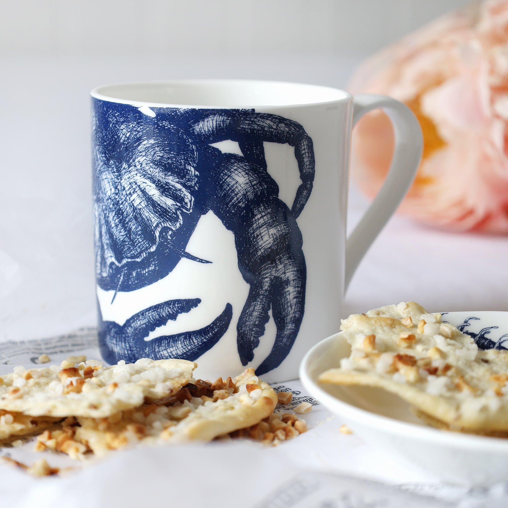 White mug with dark blue crab illustration sitting on a table with a plate & biscuits and flowers in the background.