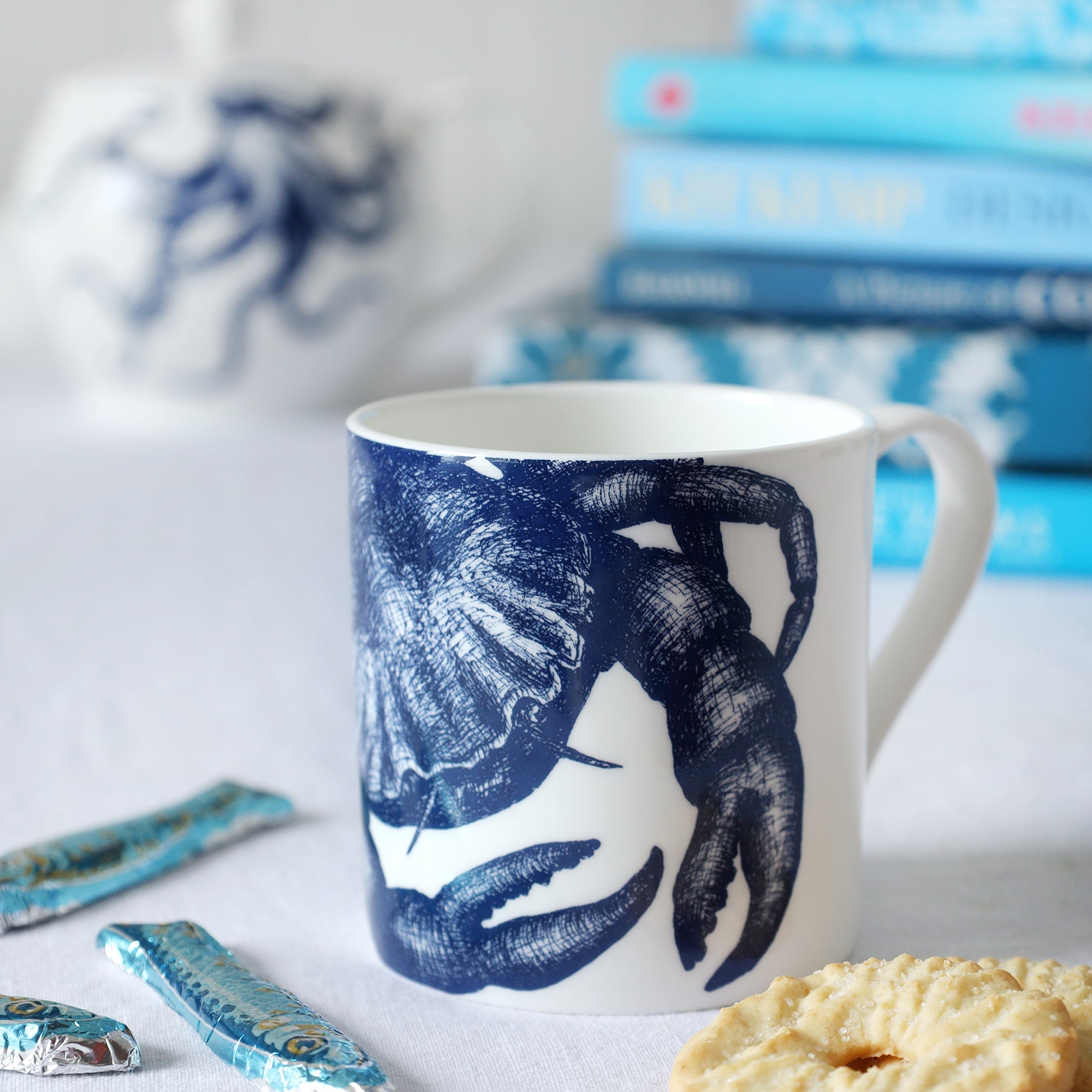 Close up of a white bone china mug with a blue illustrated crab on it, sitting on a table with biscuits and sardine chocolates and a pile of out of focus blue books and teapot in the background. 