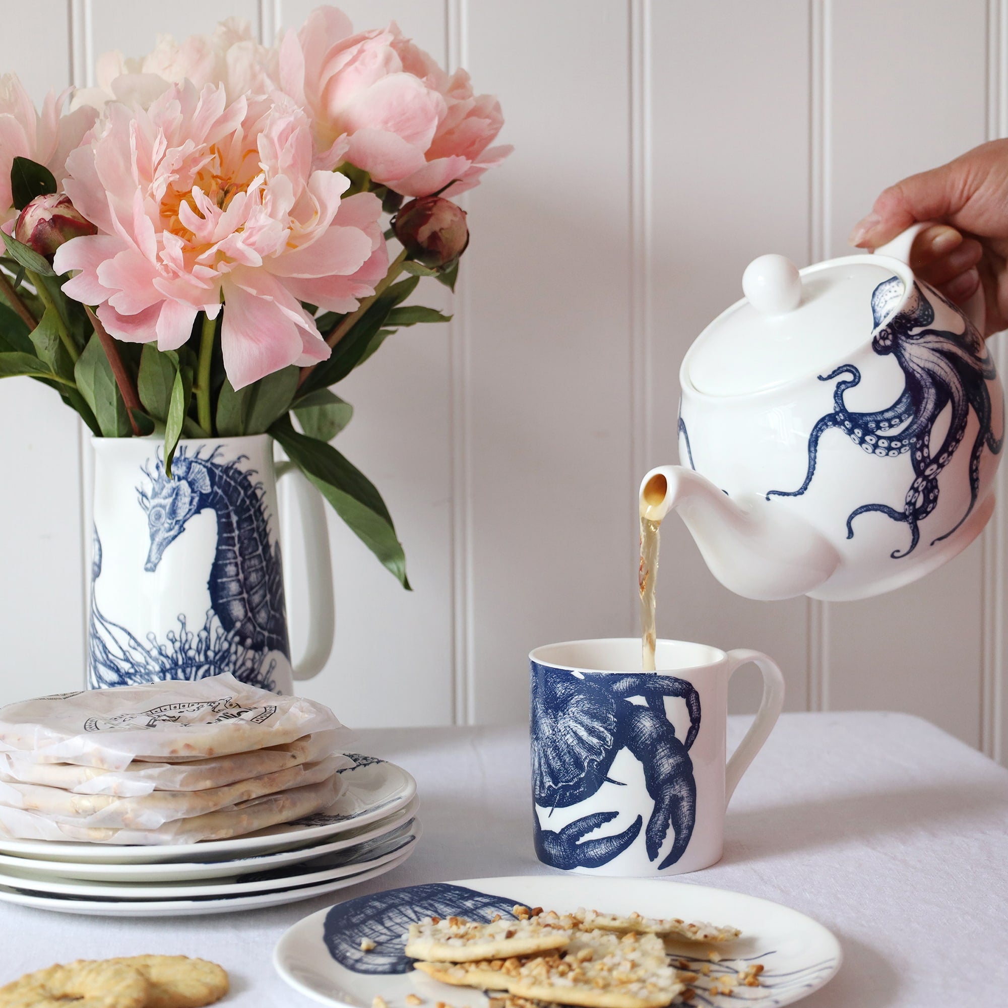 A blue & white informal; table setting set against a white shiplap wall and linen tablecloth. An octopus design adorns a teapot which is pouring tea into a white bone china mug decorated with a blue illustrated crab.
