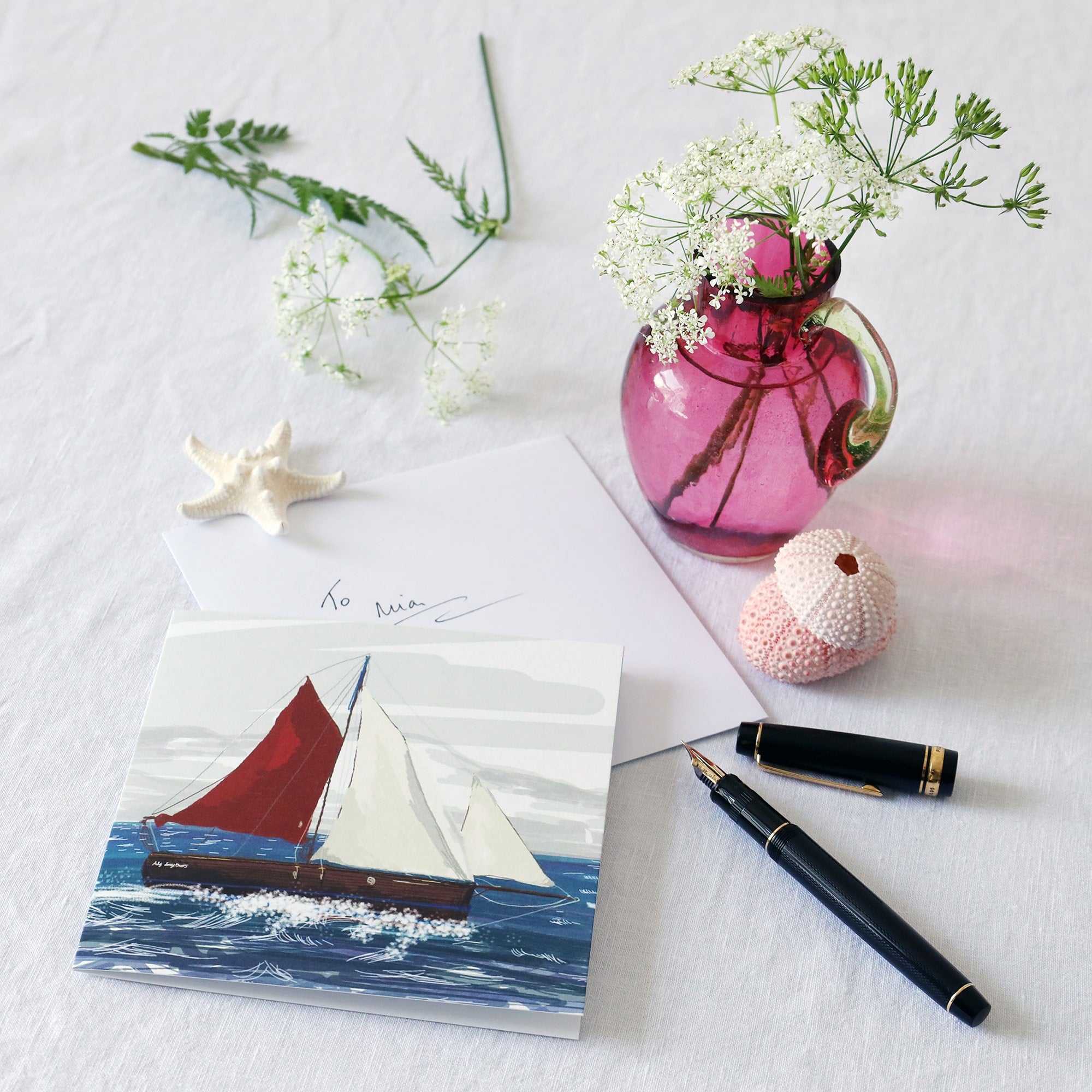 greeting card with illustration of a cornish crabber boat sailing in the sea lying on a white table cloth with a fountain pen, hand written envelope shells and a small cranberry glass jug with wild flowers in