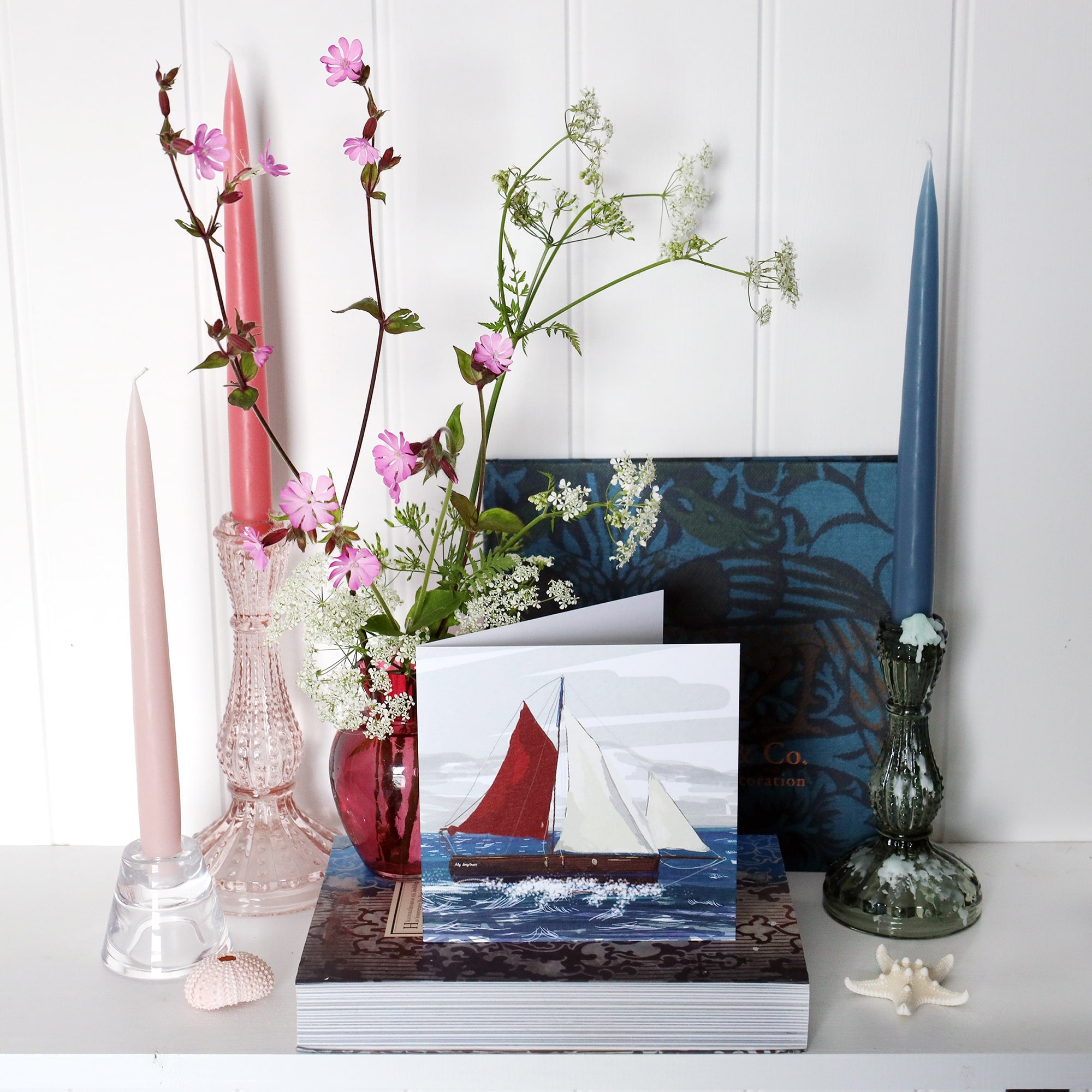 greeting card with illustration of a cornish crabber boat sailing in the sea on shelf with pink and blue candles in candlesticks and a small cranberry glass jug with wild flowers in