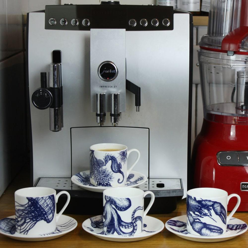 Four Bone China white espresso cups in hand drawn illustrations in our Octopus,Seahorse,Jellyfish and the Lobster designs in Navy with matching saucers in front of large coffee machine next to a red food processor