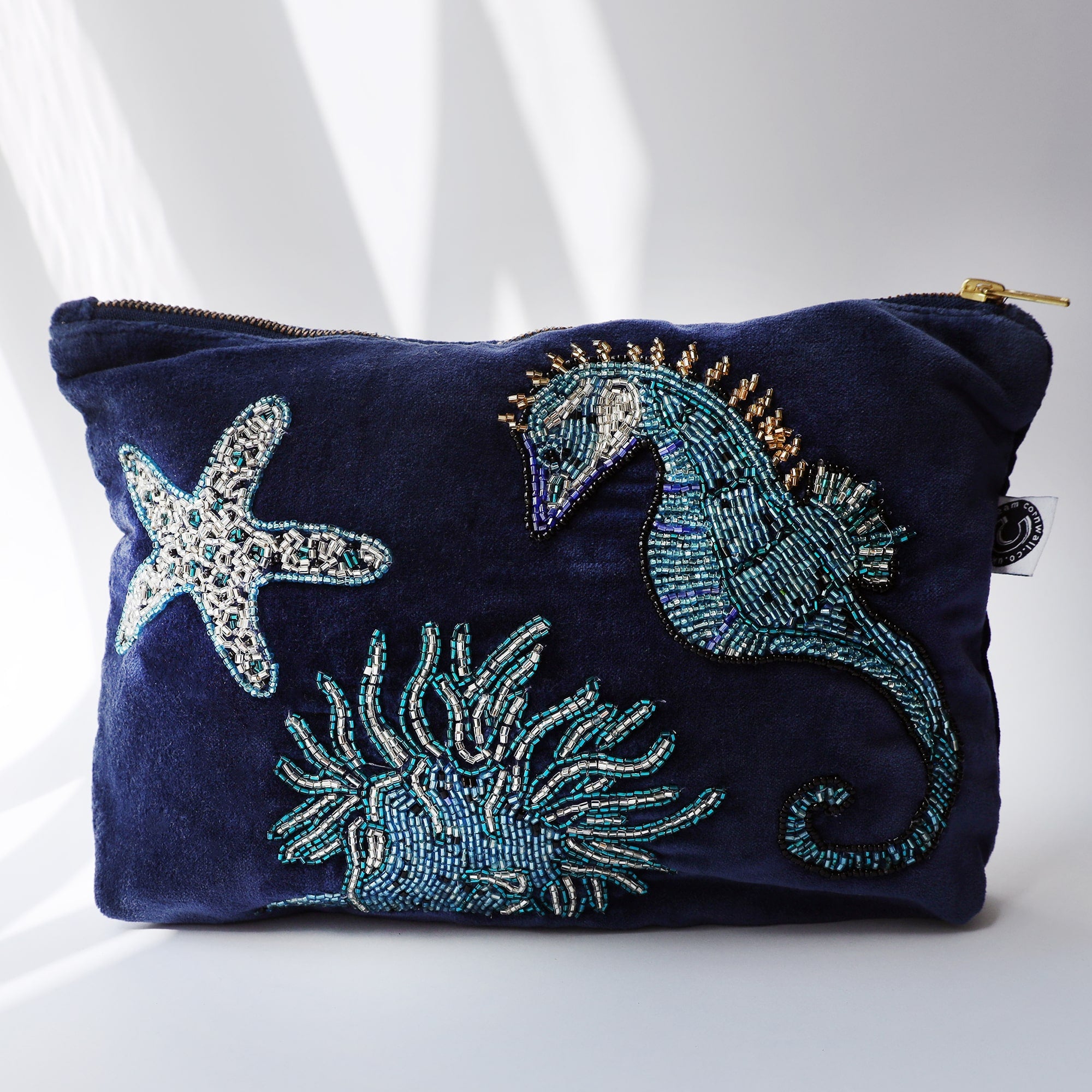 A navy velvet pouche decorated with a beaded seahorse, anemone and starfish on a white background with the su casting shadows across it.