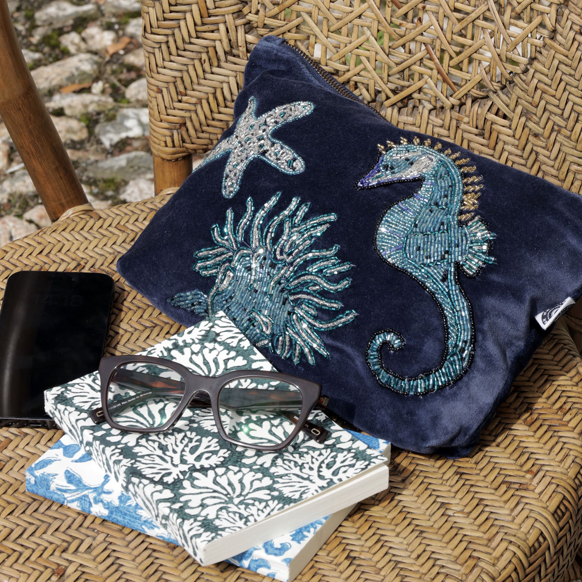 A navy velvet pouche decorated with a beaded seahorse, anemone and starfish on a wicker chair with mobile phone, notebooks and a pair of glasses.