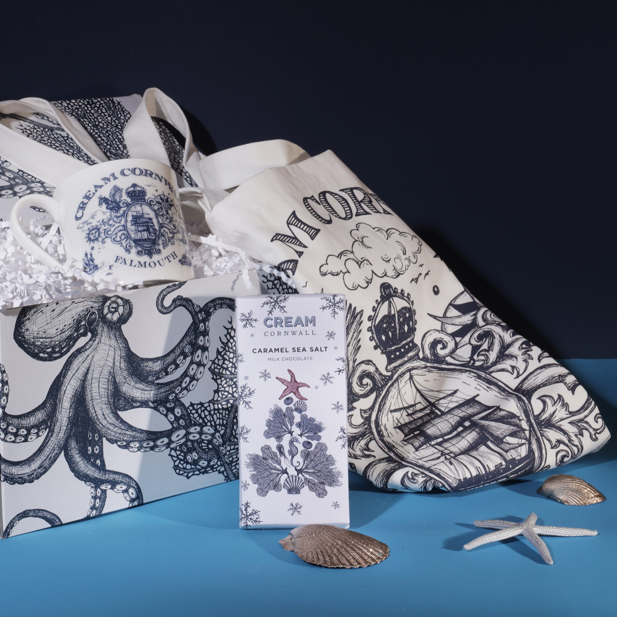 A Cream Cornwall Falmouth-themed gift set featuring a mug, tote bag, and a bar of caramel sea salt chocolate. The mug and tote bag are decorated with intricate nautical illustrations in blue, including ships and maritime symbols. The items are displayed in a box adorned with octopus illustrations, and decorative seashells and starfish are arranged around the set, enhancing the coastal theme against a dark blue background.