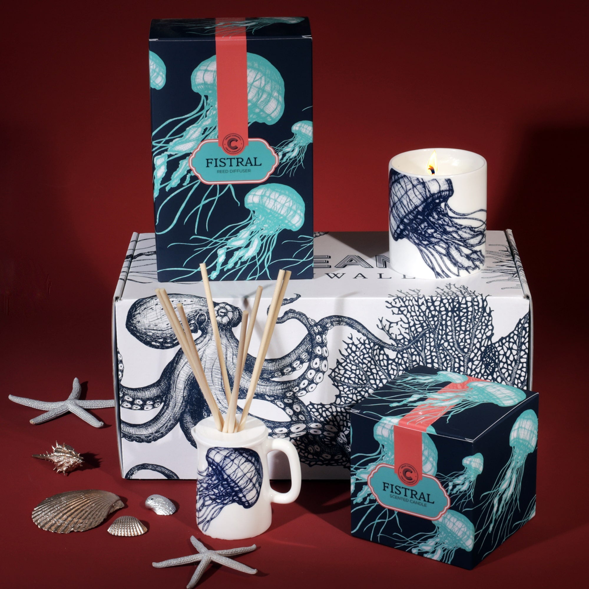 A Cream Cornwall Fistral-themed fragrance gift set, featuring a reed diffuser and a scented candle. The packaging and candle holder are adorned with jellyfish illustrations in teal and navy. The products are displayed on a box decorated with octopus and coral designs, accompanied by decorative seashells and starfish, enhancing the coastal theme against a deep red background.