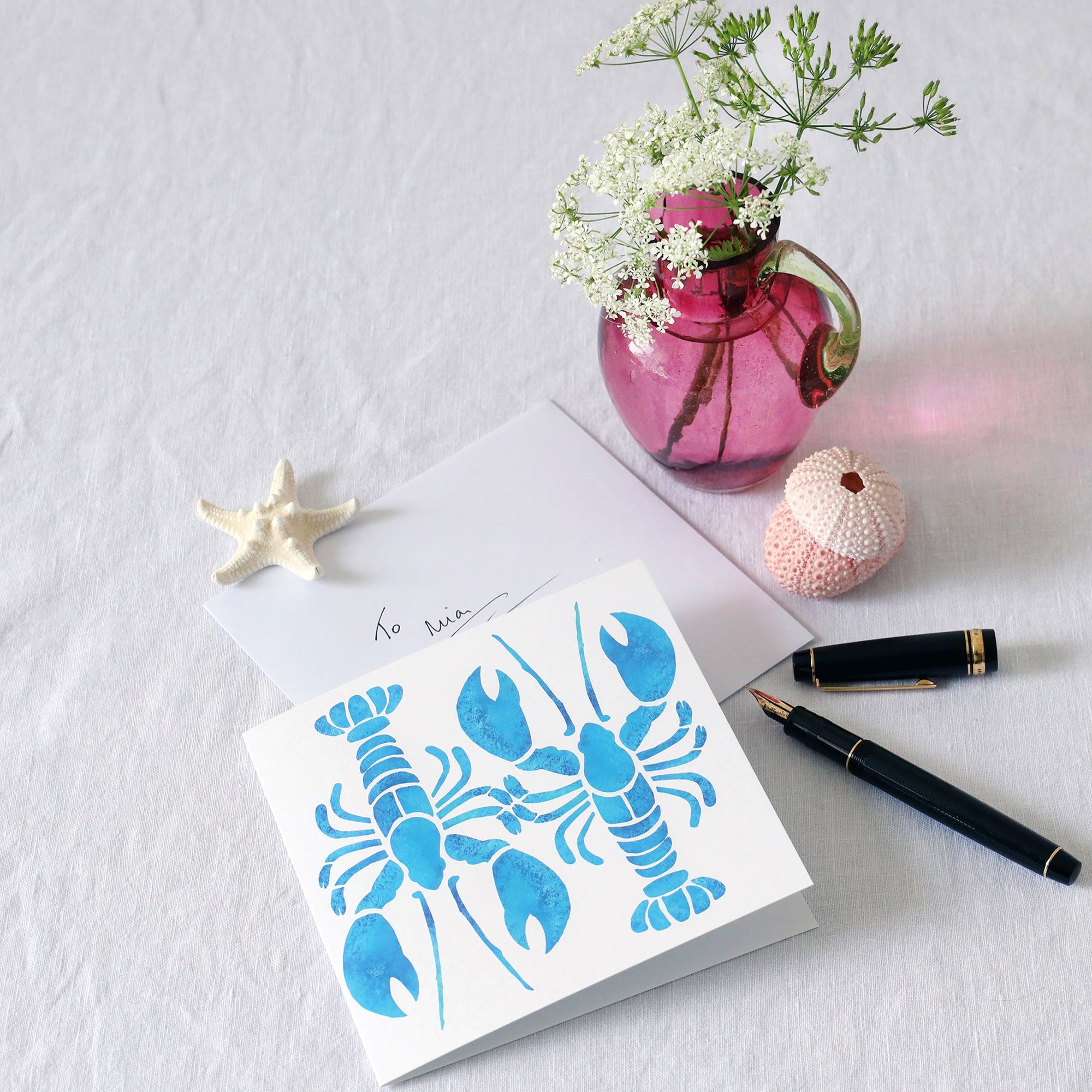 greeting card with 2 turquoise illustrated lobsters one facing up and the other facing down, abstract and filled with a mottled blue effect on a white background lying on a white table cloth with a fountain pen, hand written envelope shells and a small cranberry glass jug with wild flowers in