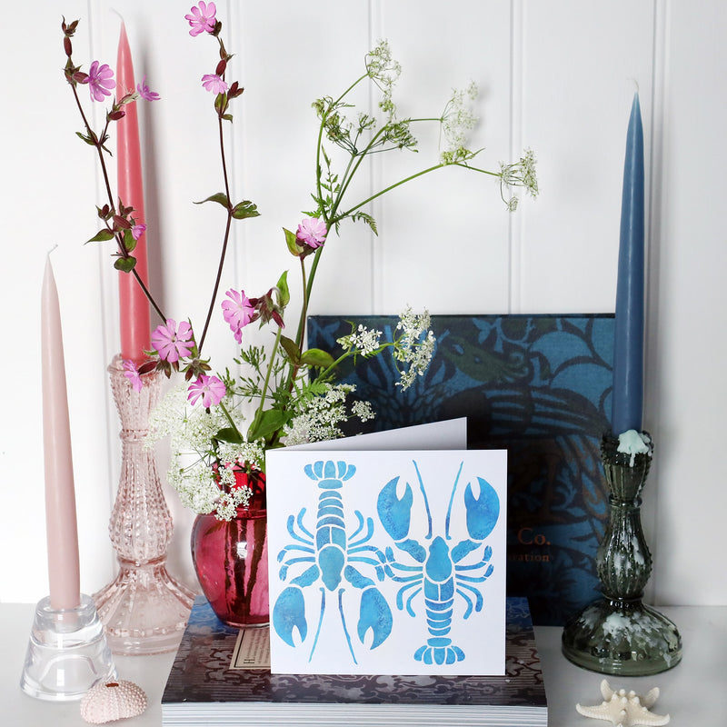 greeting card with 2 turquoise illustrated lobsters one facing up and the other facing down, abstract and filled with a mottled blue effect on a white background on shelf with pink and blue candles in candlesticks and a small cranberry glass jug with wild flowers in