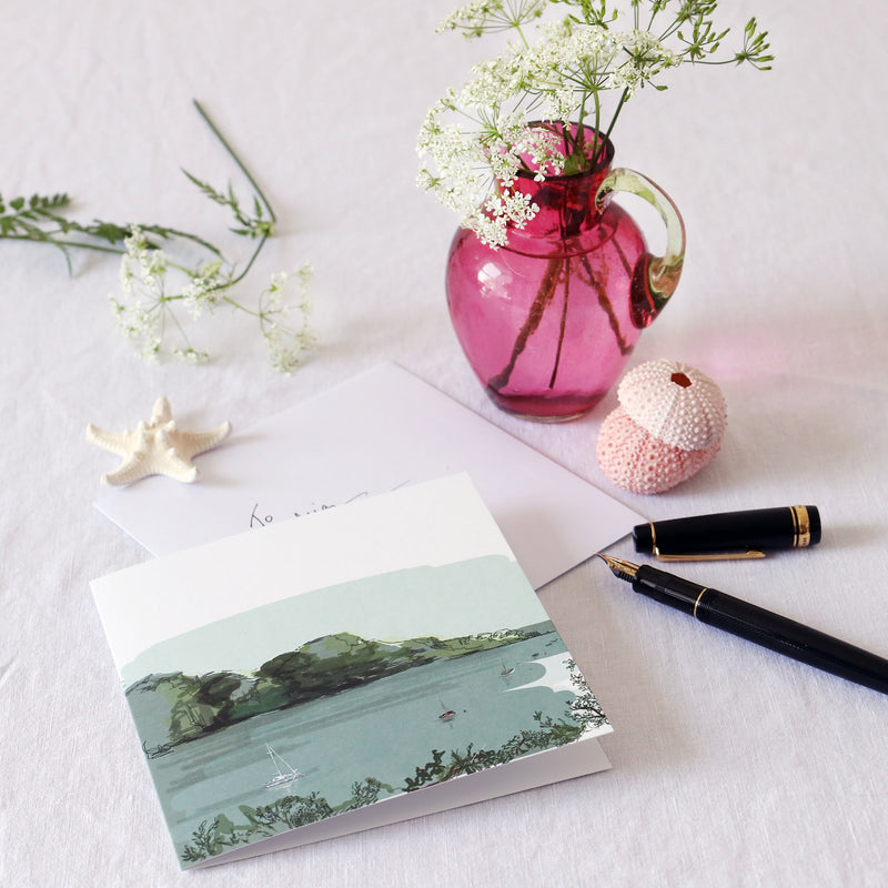 greeting card with illustration of frenchmans creek with small boats sailing in the water in muted blue and green tones  lying on a white table cloth with a fountain pen, hand written envelope shells and a small cranberry glass jug with wild flowers in 