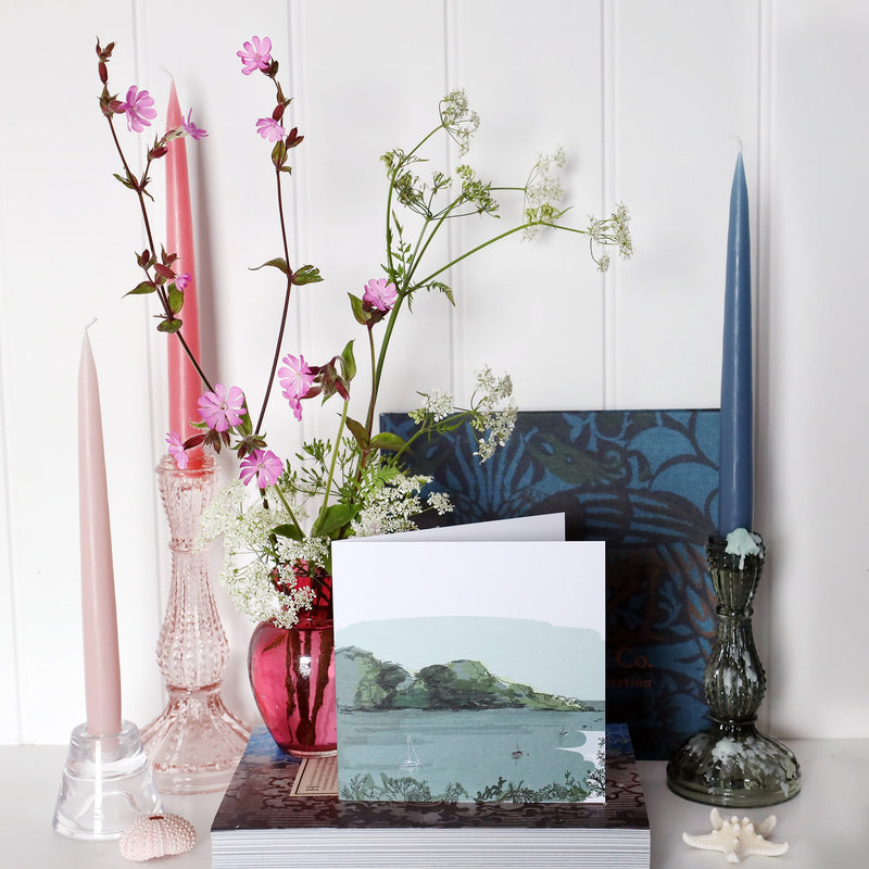 greeting card with illustration of frenchmans creek with small boats sailing in the water in muted blue and green tones  on shelf with pink and blue candles in candlesticks and a small cranberry glass jug with wild flowers in