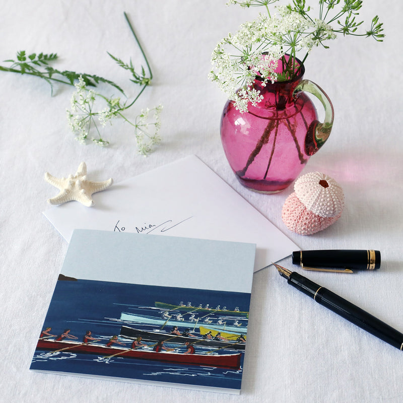 greeting card with illustration of 5 gig boats racing across a blue sea lying on a white table cloth with a fountain pen, hand written envelope shells and a small cranberry glass jug with wild flowers in 