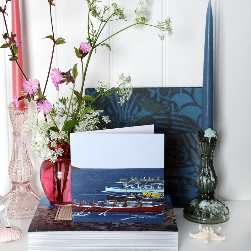 greeting card with illustration of 5 gig boats racing across a blue sea on shelf with pink and blue candles in candlesticks and a small cranberry glass jug with wild flowers in